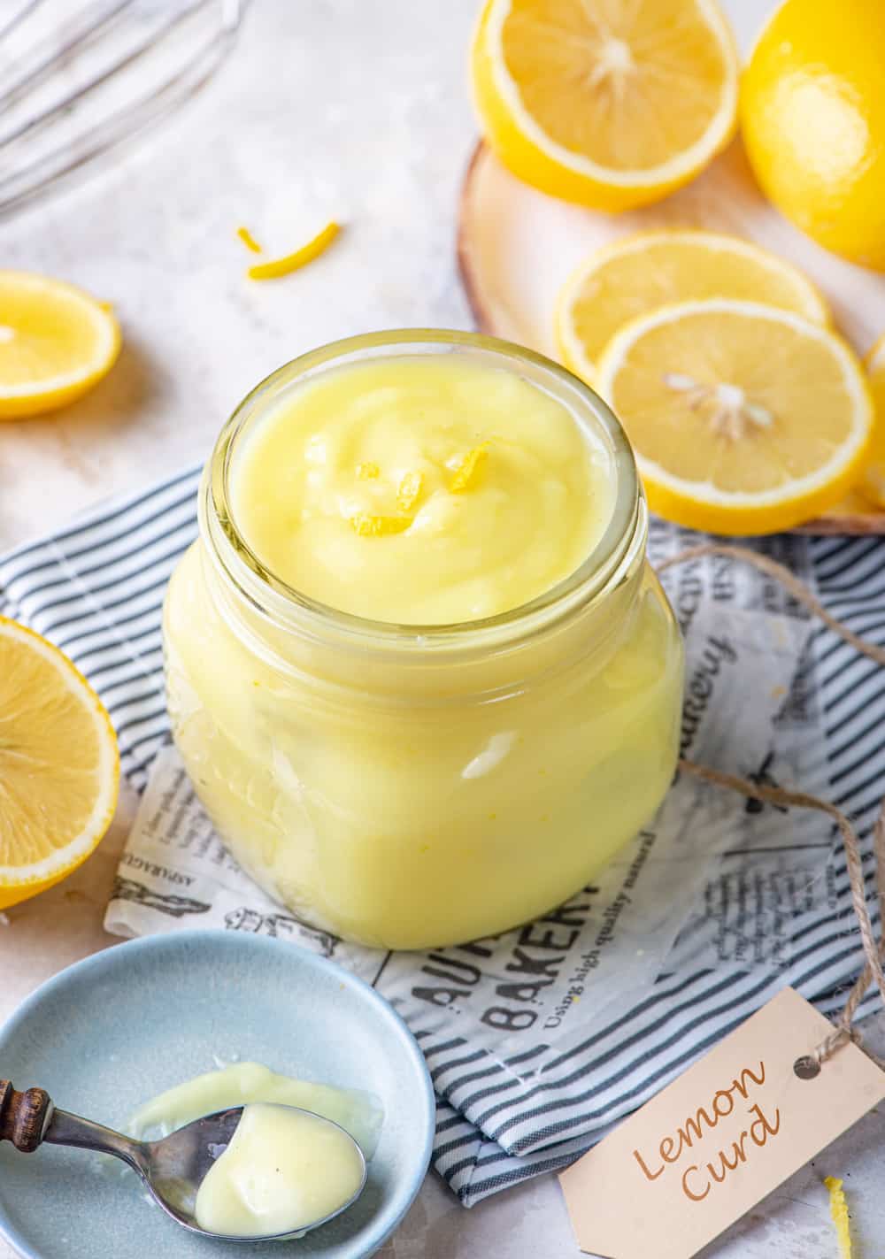 A glass jar full of vegan lemon curd. The jar is set on a piece of white newspaper that is on a white and blue stripped tablecloth. A plate of sliced lemons is behind the jar to the left and a small blue dish with a spoon full of vegan lemon curd on top of it. A brown tag that says lemon curd is to the right of the bowl.