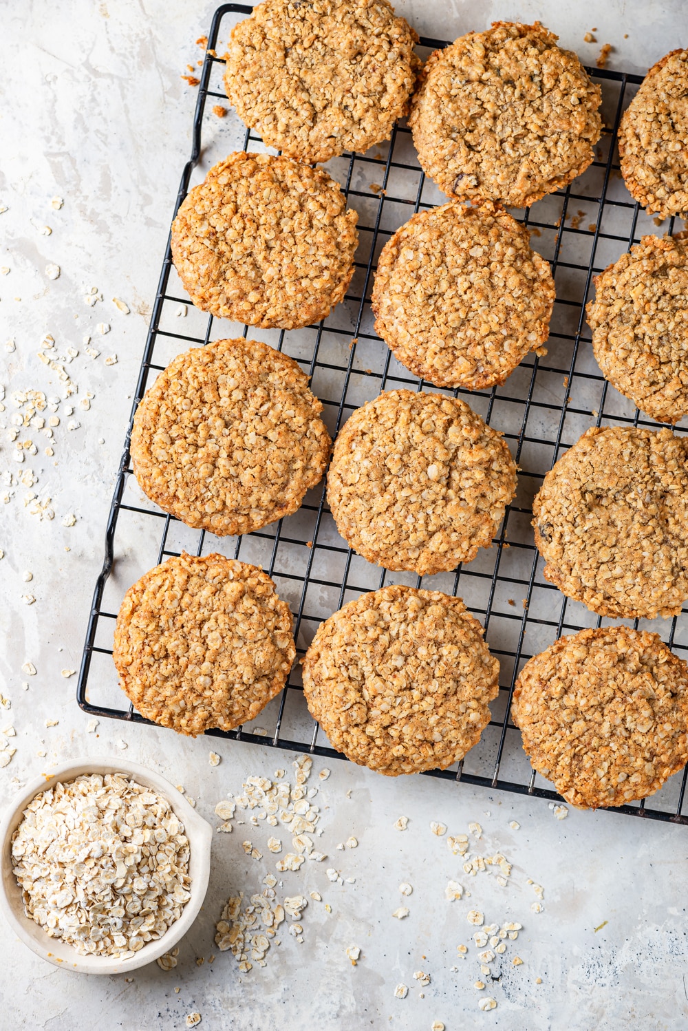 An overhead view of three rows of four vegan oatmeal cookies sitting on a black wire rack. A small white bowl of oats is set in front of the wire rack, with a few oats on scattered on the white counter everything is sitting on.