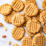 About fourteen vegan peanut butter cookies scattered on a white counter. Some cookies are laying flat and others are overlapping one another. There is a spoonful of peanut butter at the bottom of all the cookies and few peanuts scattered on the outsides of the group of cookies.