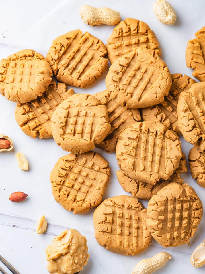 About fourteen vegan peanut butter cookies scattered on a white counter. Some cookies are laying flat and others are overlapping one another. There is a spoonful of peanut butter at the bottom of all the cookies and few peanuts scattered on the outsides of the group of cookies.