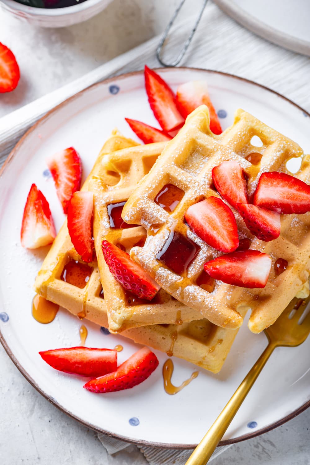 An overhead view of three square vegan waffles unevenly stacked on top of each other with maple syrup and cut up strawberries on them. The waffles are on white plate and there are a few pieces of strawberries on the plate as well. A gold fork is at the front of the plate with the tip of the prongs hidden by the top waffle. The plate is on a tablecloth on a white counter.