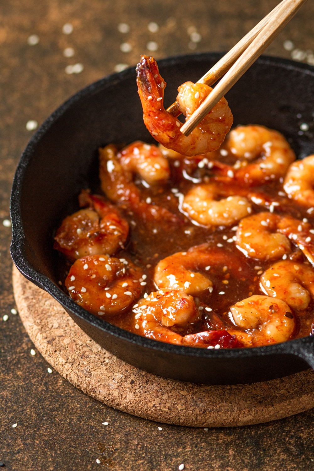 Several pieces of shrimp in a fire cracker sauce in a skillet. There is a piece of shrimp in between chopsticks being held above the skillet. The skillet is on a circular cork board on a brown counter.