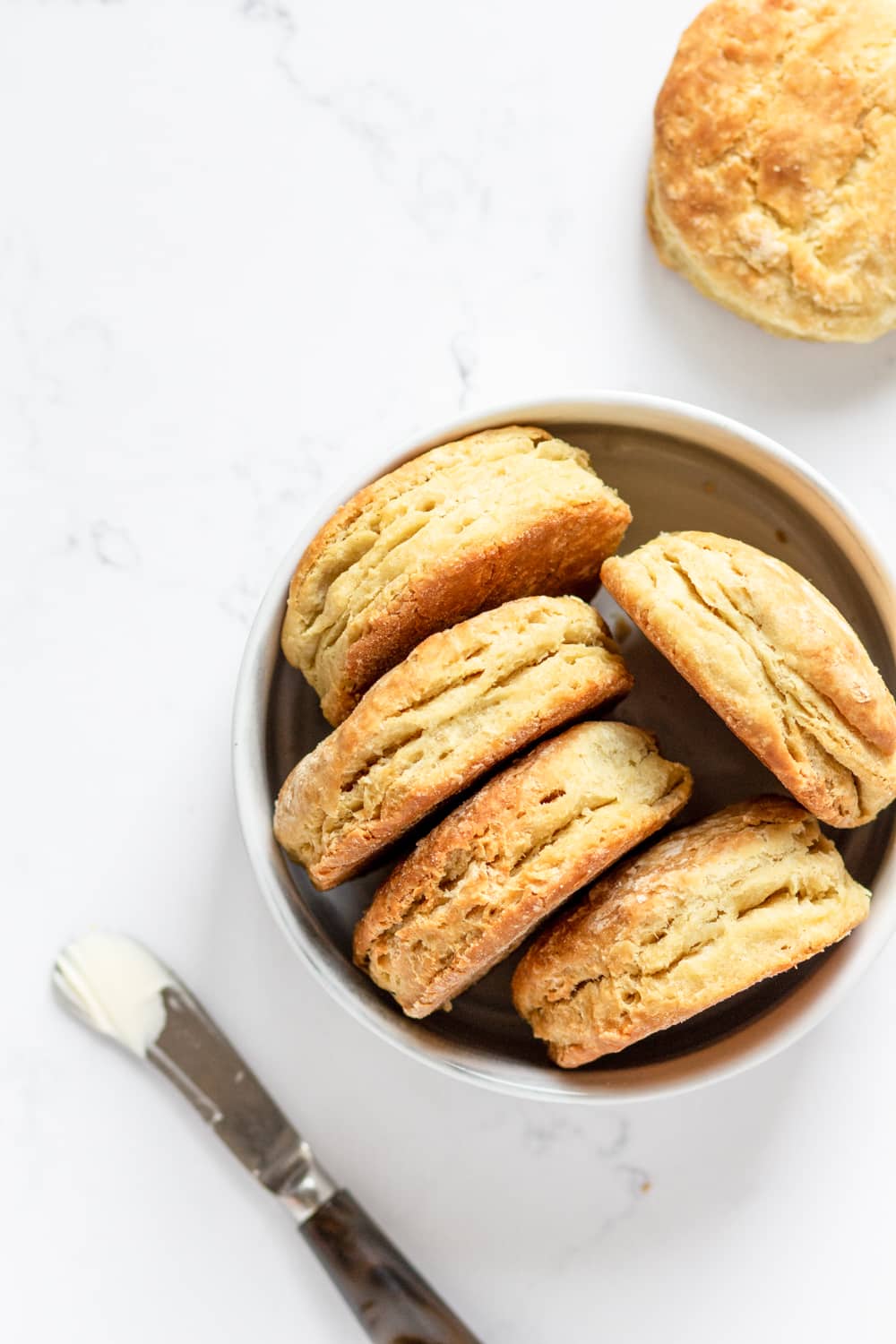 An overhead view of five vegan biscuits on their side in a white bowl. There is a knife with butter on it in front of the bowl. A vegan biscuit is behind the bowl. Everything is on a white counter.