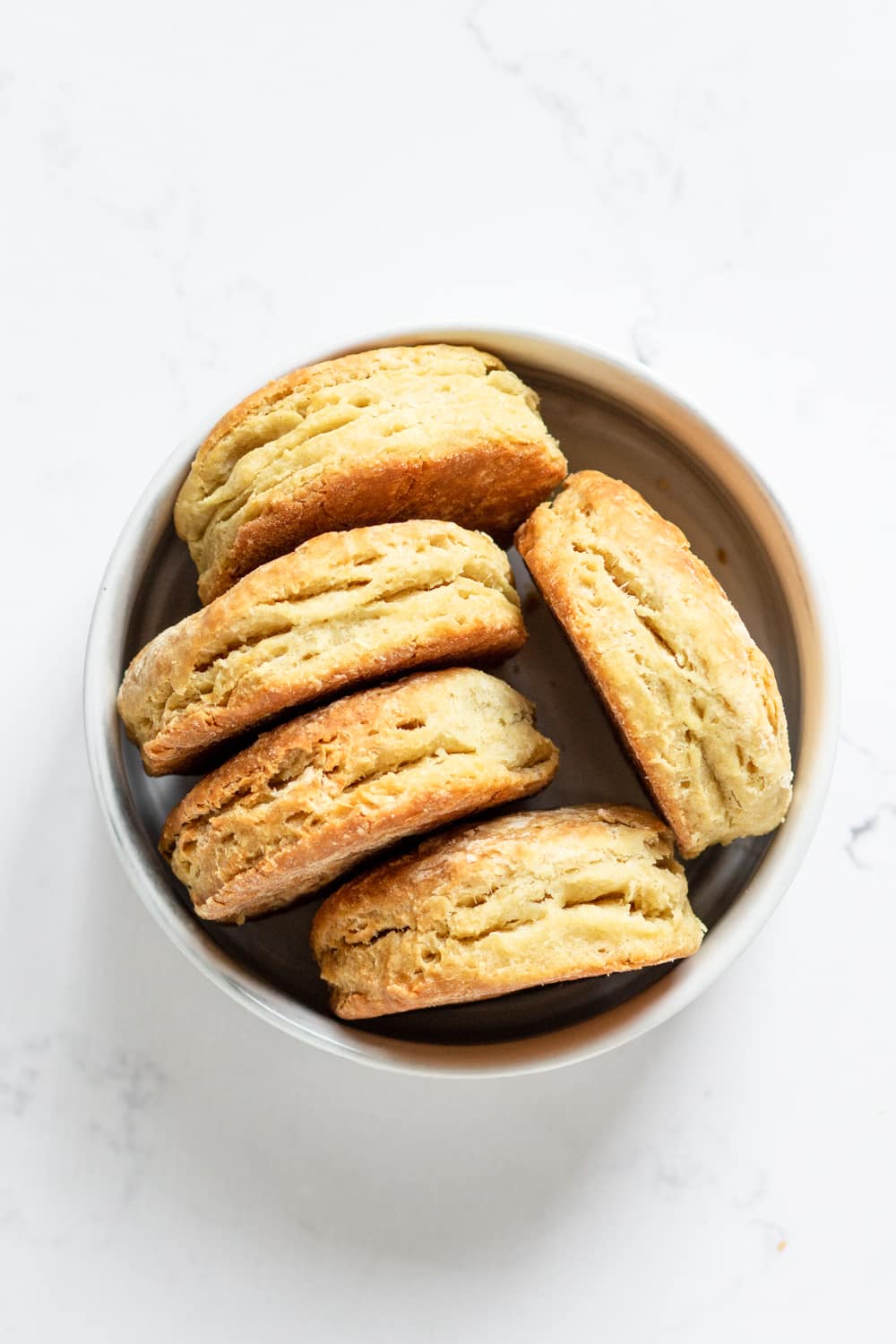 An overhead view of a white bowl filled with five vegan biscuits on their side. The bowl is on a white counter.