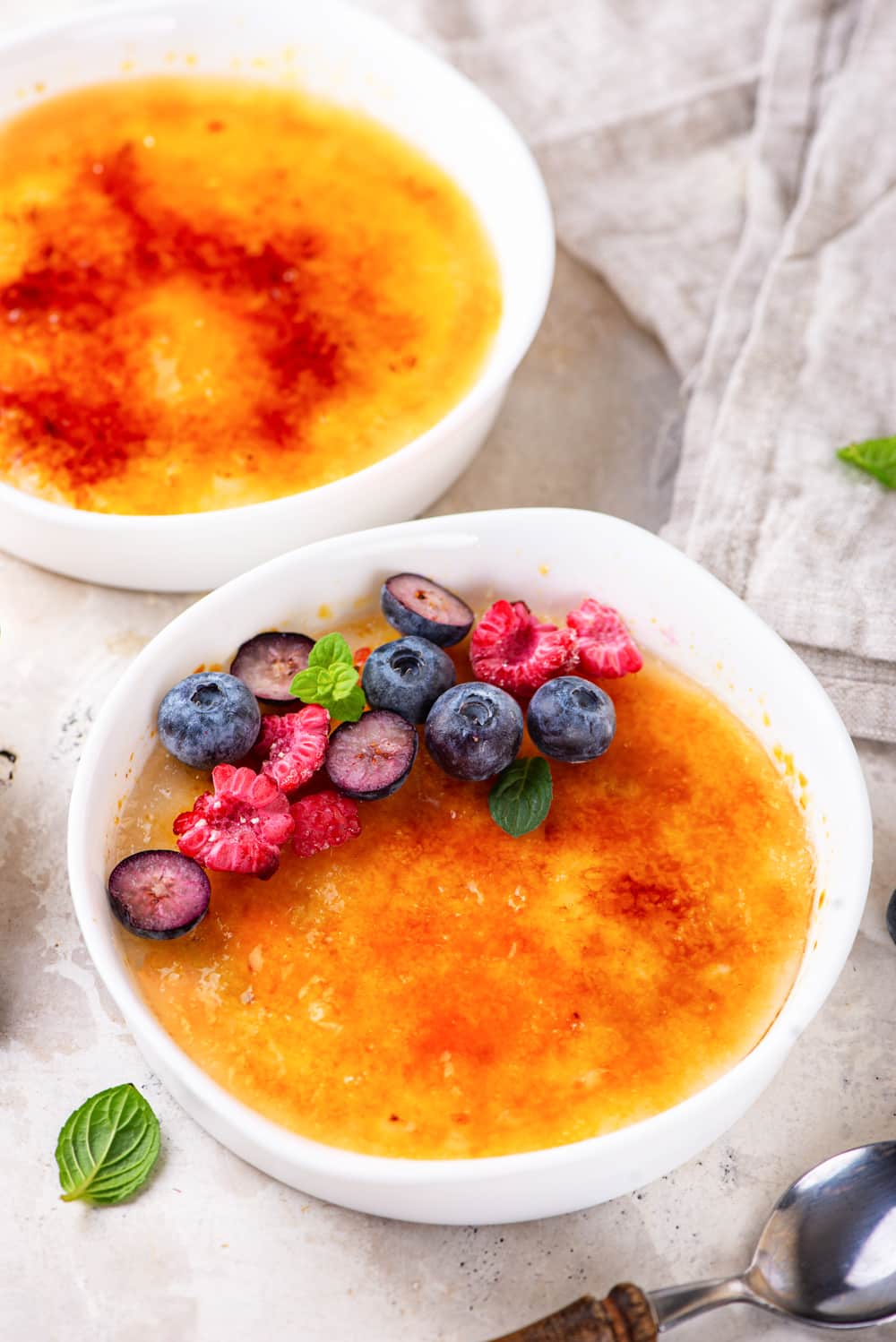One ramekin filled with creme brulee, with sliced raspberries and blueberries on the back third of the surface of the creme brulee. A ramekin filled with creme brulee is behind it. Both ramekins are on a white counter.