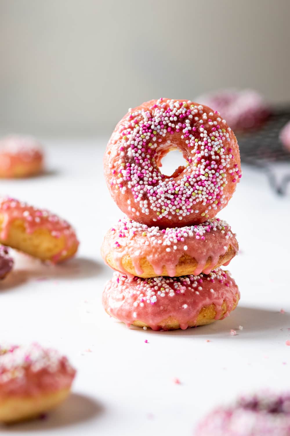 Two vegan donuts stacked on top of each other with a third donut on its side on top of them. The donuts have pink glaze with white, pink, and yellow ball sprinkles on top. The stack of donuts is on a white counter.