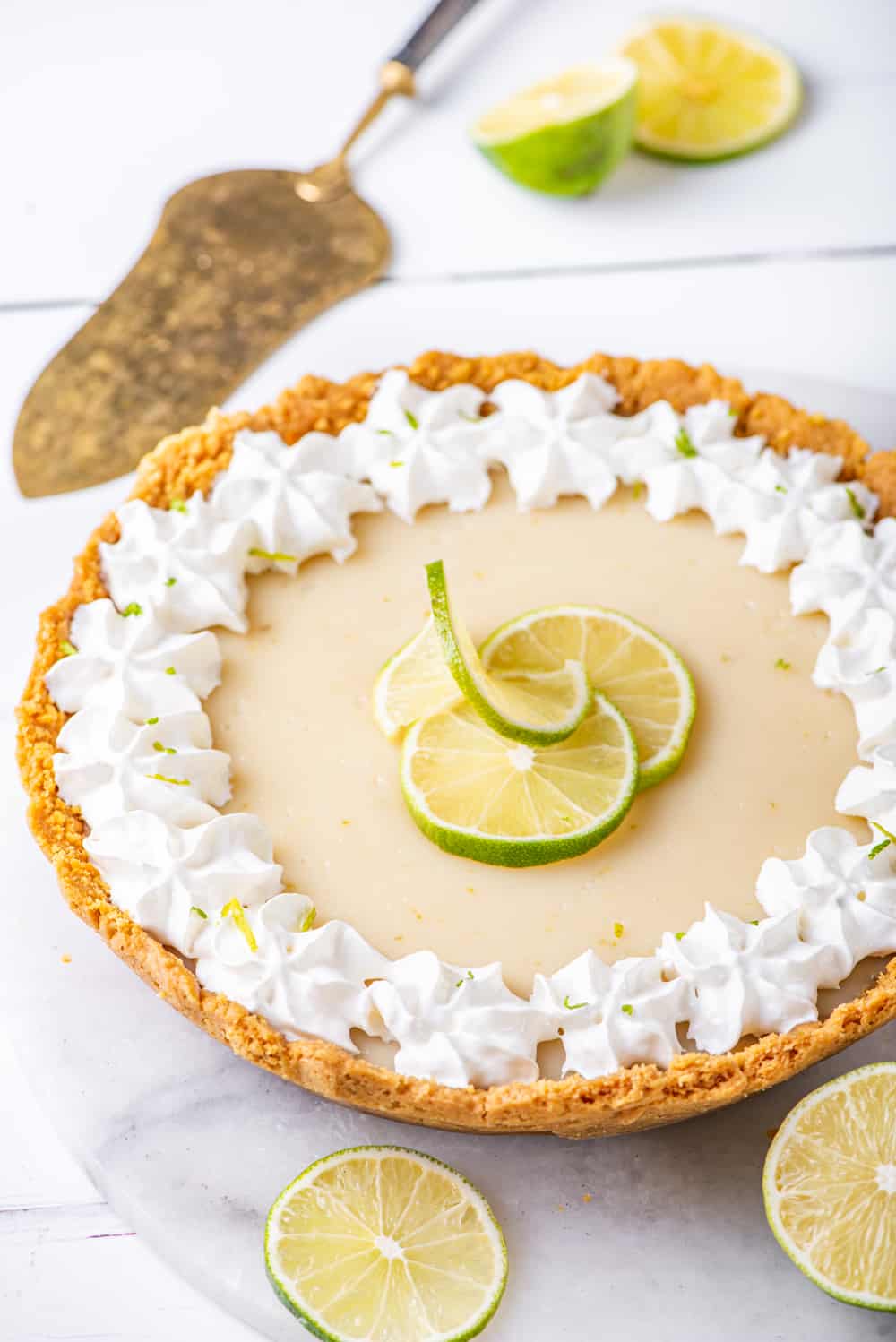 A whole vegan key lime pie on a white marble board. A pie server is behind the pie and a few slices of limes are behind it and in front of the pie. Everything is on a white counter.