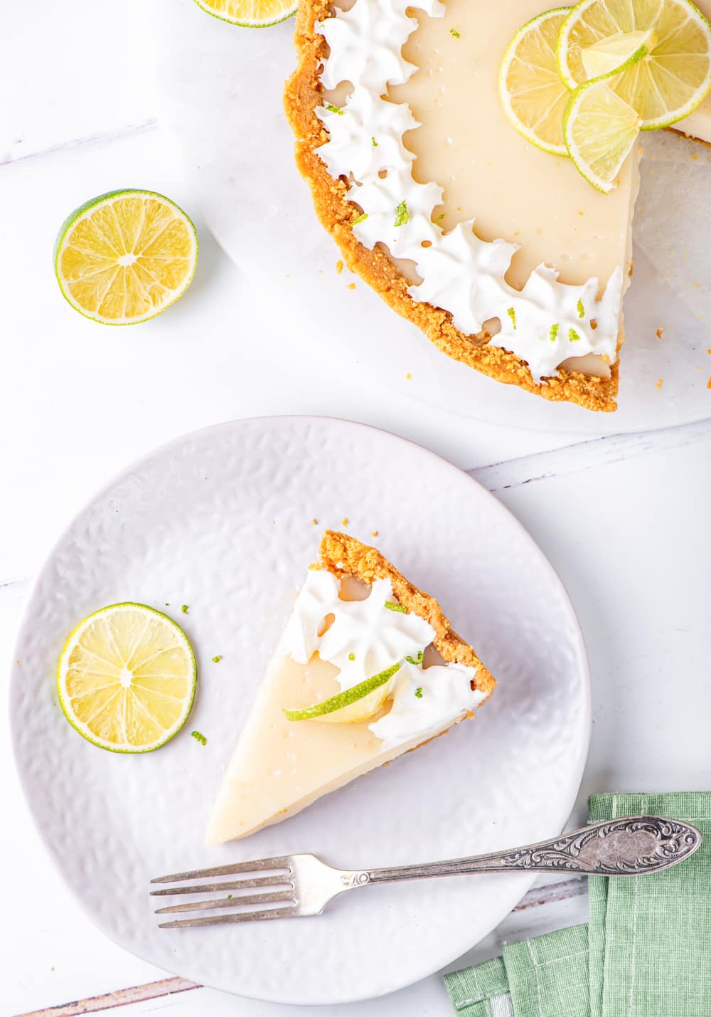 An overhead view of a piece of vegan key lime pie on a whit plate. A lime slice and metal fork are on the plate at the sides of the pie. Part of the vegan key lime pie is set behind the plate, showing where the piece was sliced out of. One lime slice is between the pie and plate and everything is on a white counter.