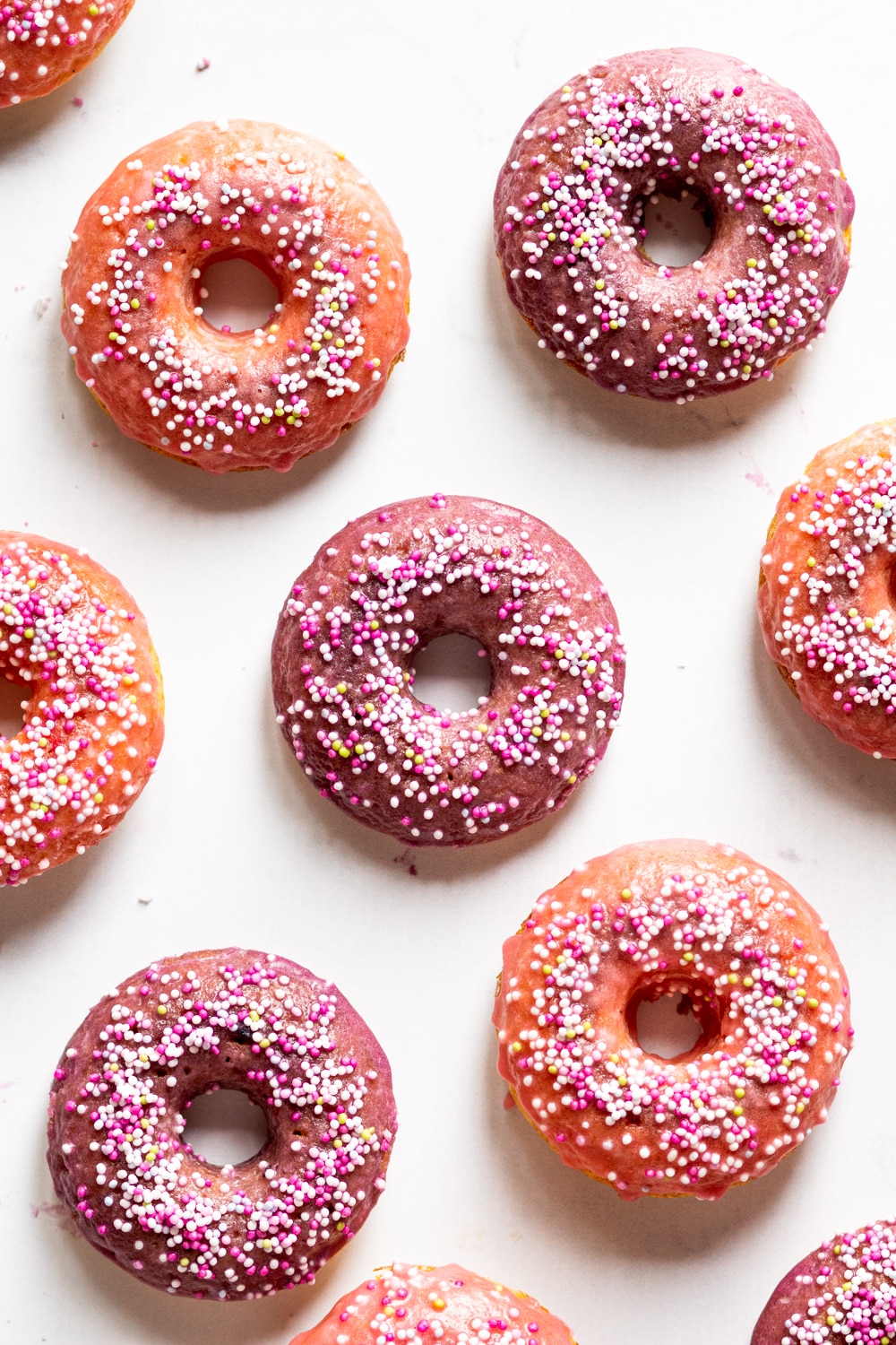 An overhead view of five whole vegan donuts with four halves of vegan donuts on the sides. The donuts all have a pink glaze with white, pink, and yellow ball sprinkles on top.