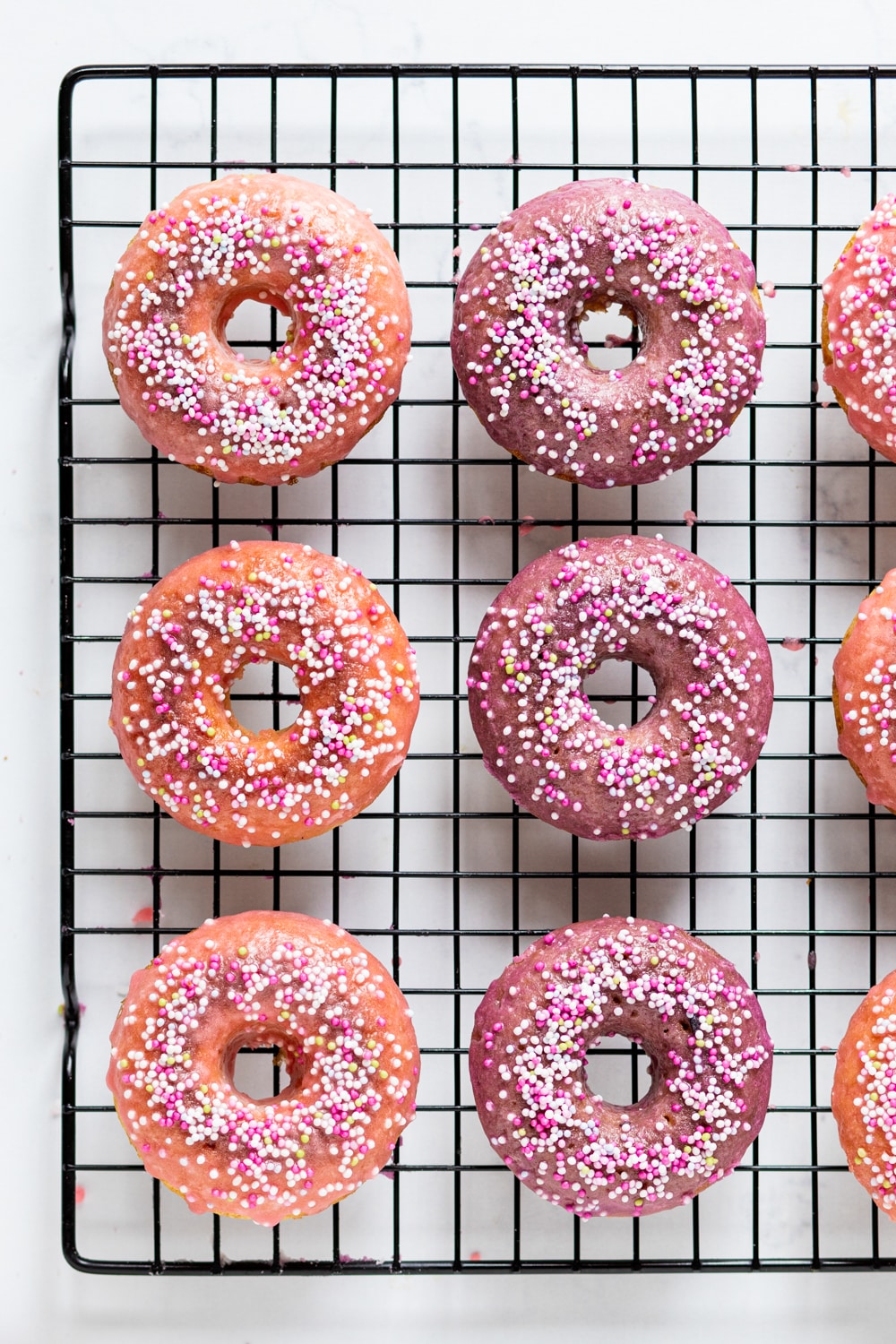 An overhead view of two rows of vegan donuts with three donuts in each row on a black wire rack. There is pink glaze with white, pink, and yellow ball sprinkles on top.