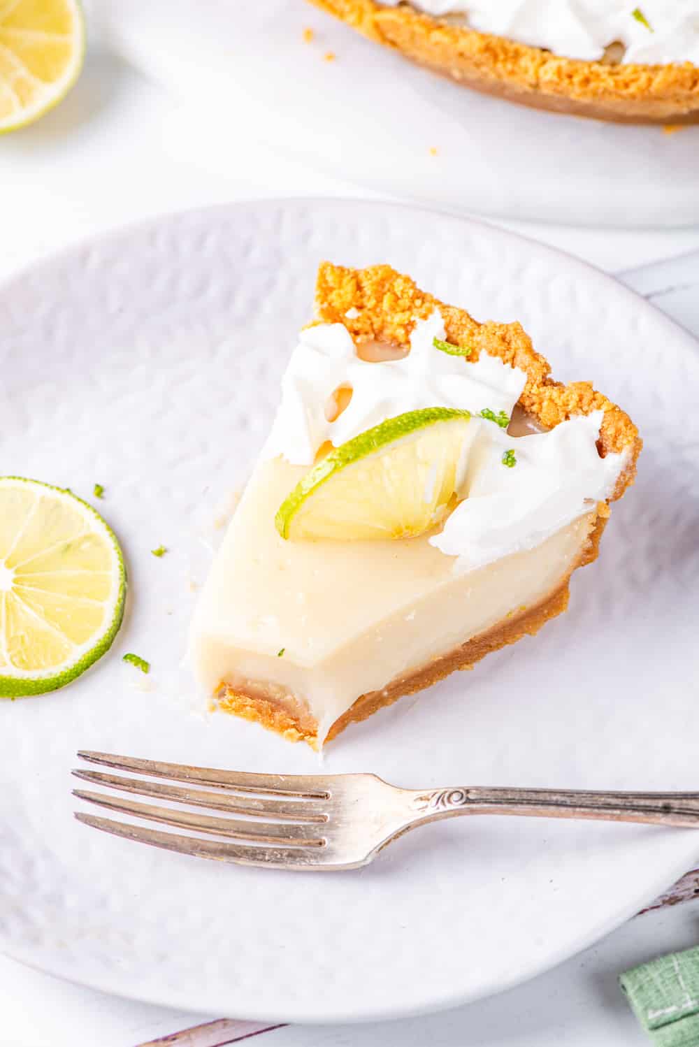 A piece of vegan key lime pie with the tip sliced off and a metal fork in front of it, all on a white plate. The plate is on a white counter.