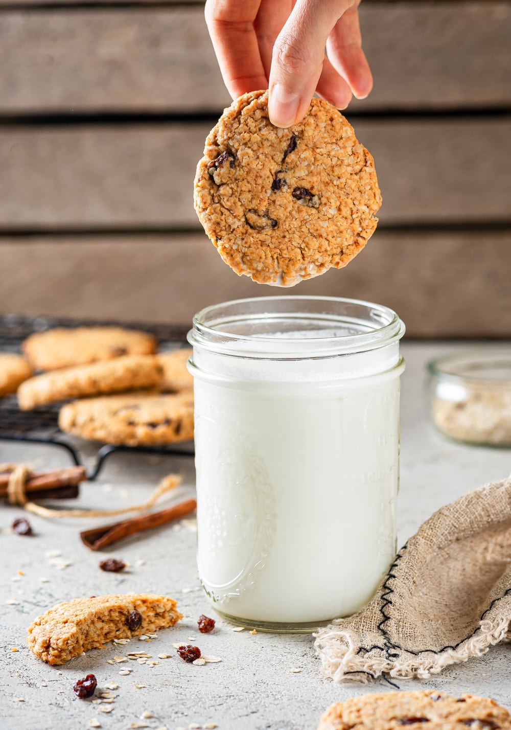 One vegan oatmeal raisin cookie being lowered down by a hand, just hovering above a glass jar of milk. A few cookies are on a black wire rack set behind and the to the left of the jar of milk. One half of a broken cookie is next to the jar of milk to the left and a yarn towel is to the right.