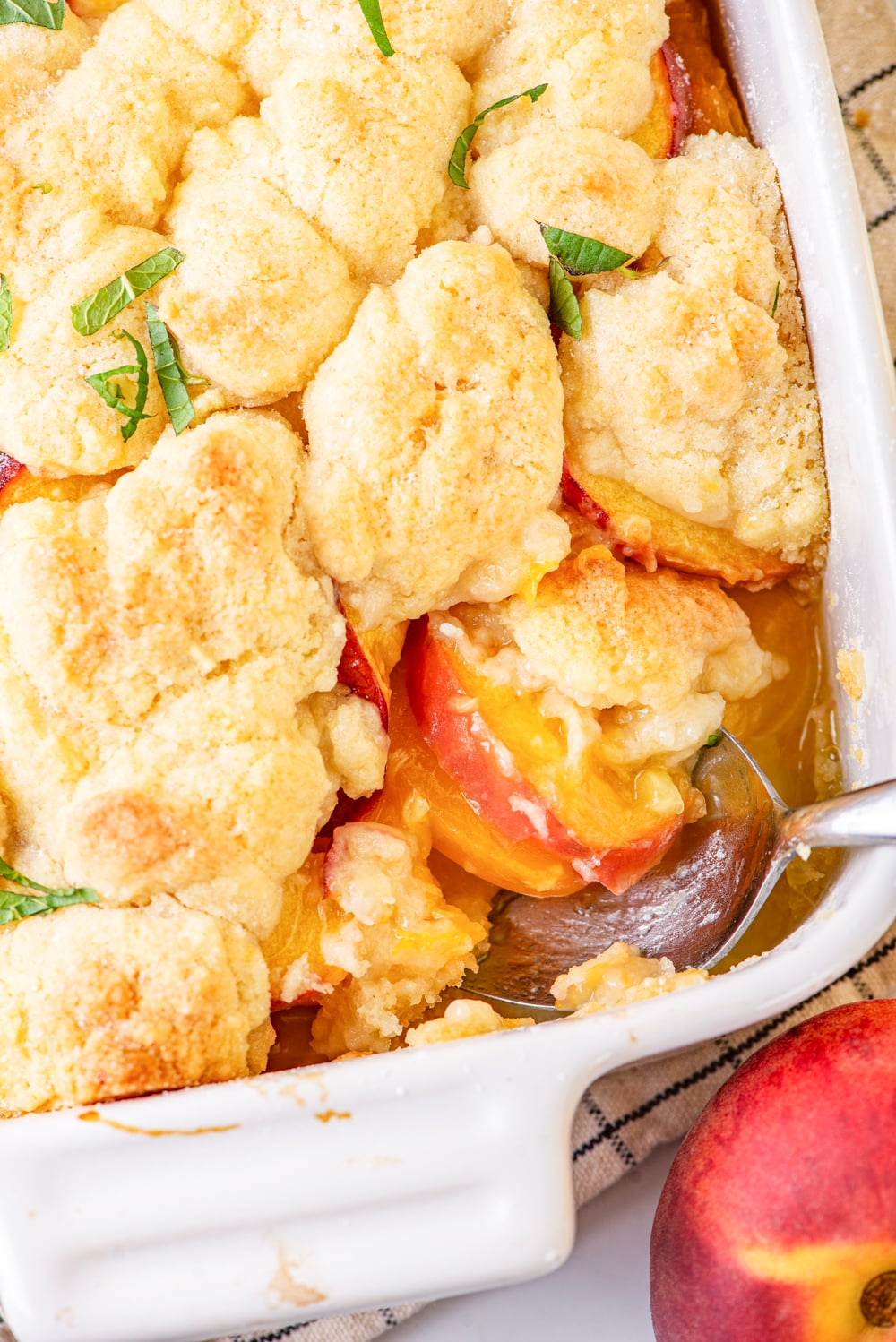 The corner of a white baking dish filled with vegan peach cobbler. There is a metal spoon head scooping the peach cobbler at the edge of the baking dish. A peach is right in front of the corner of the dish.