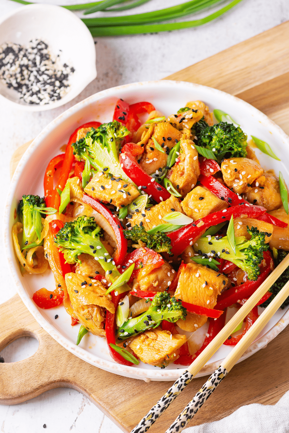 A white bowl filled with stir fry. The bowl has chicken, broccoli, and red peppers in it. The bowl is on a wooden cutting board on a white table.