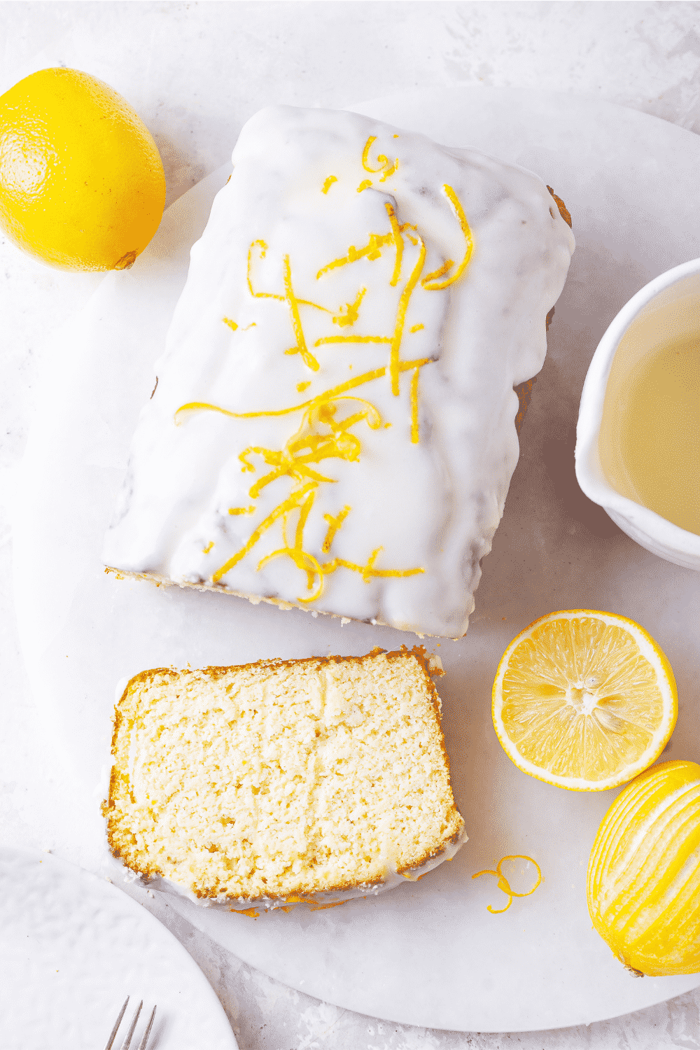 An overhead view of a loaf of keto lemon pound cake. The top of the keto cake is covered in a white keto glaze and lemon zest is on top of the glaze. A slice of keto lemon cake is in front loaf and it is flat on the white saucer. There is a sliced lemon and a whole lemon to the right of the piece of cake. A whole lemon is to the back left of the loaf.