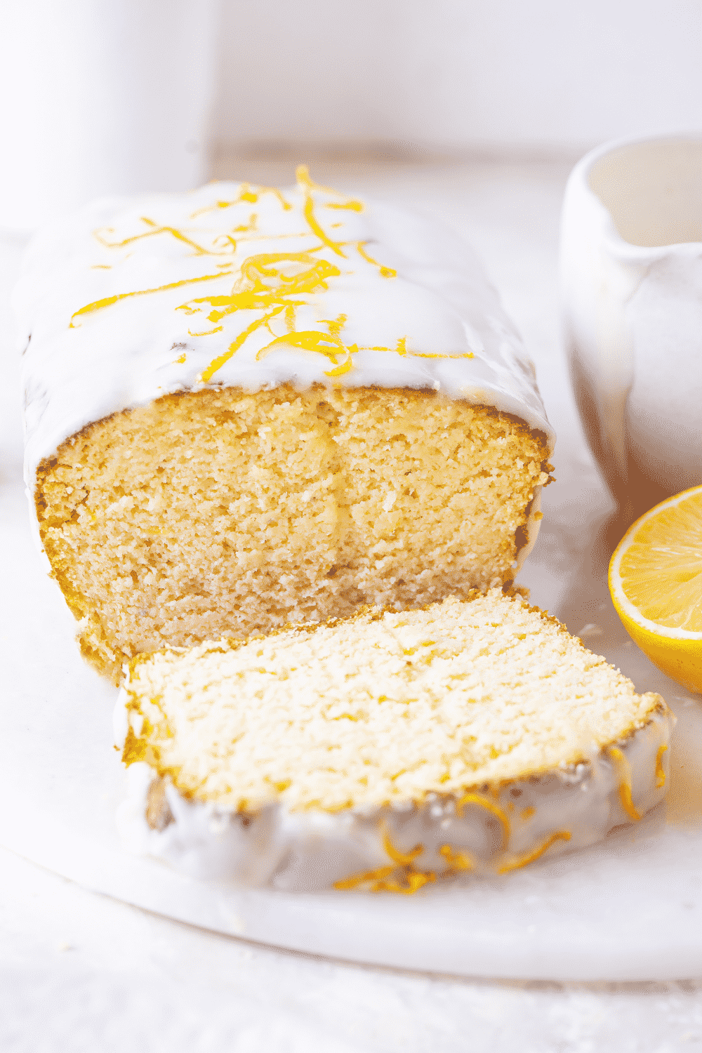A loaf of keto lemon pound cake on a white saucer. A slice off the front of loaf is lying flat directly in front of the loaf and on the saucer. There is a cup of keto glaze and a sliced lemon to the right of the loaf of keto cake.