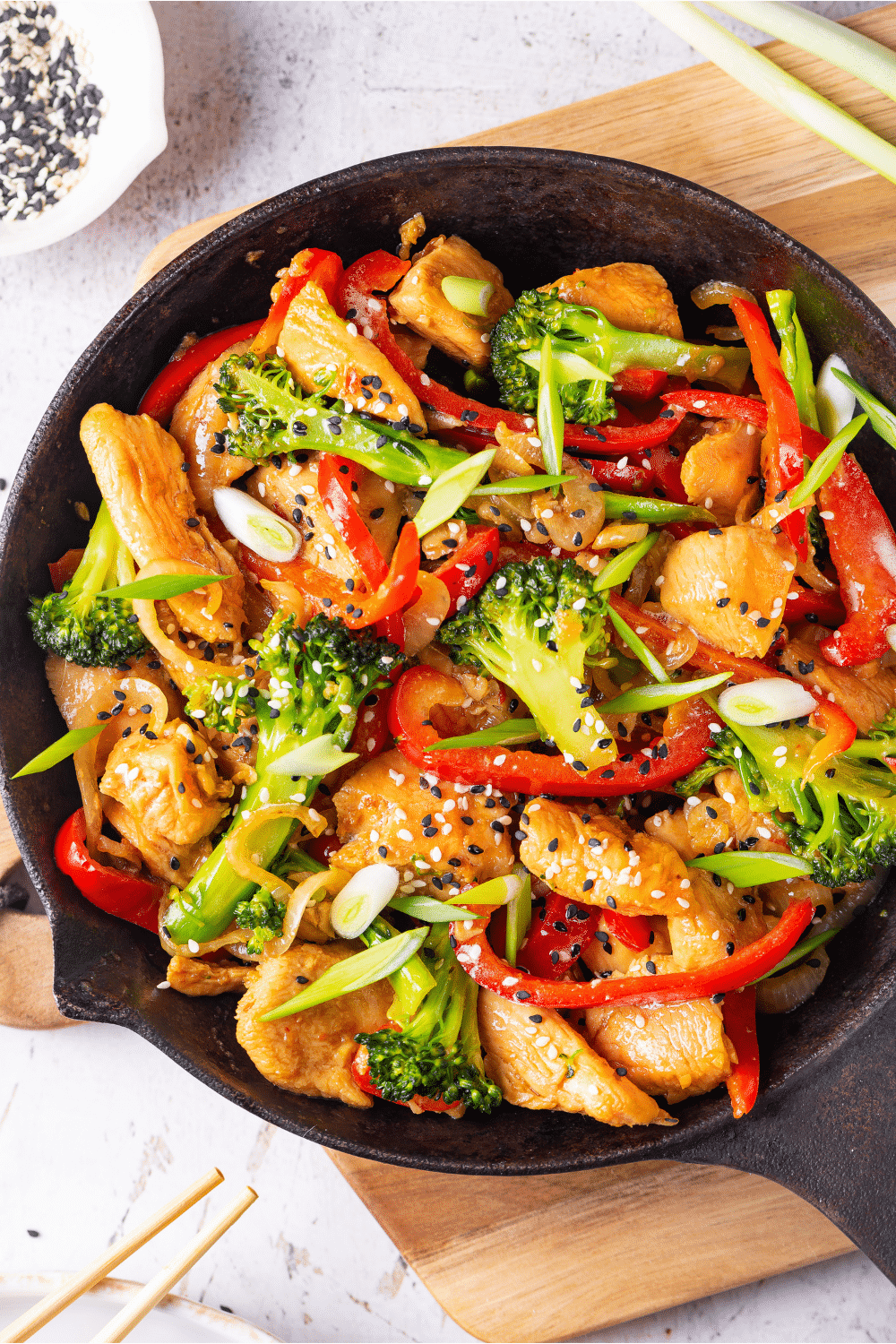 A skillet filled with keto chicken stir fry. The skillet has broccoli, red peppers, chicken, and onions in it covered in a keto stir fry sauce. The skillet is on a wooden cutting board on a white table.