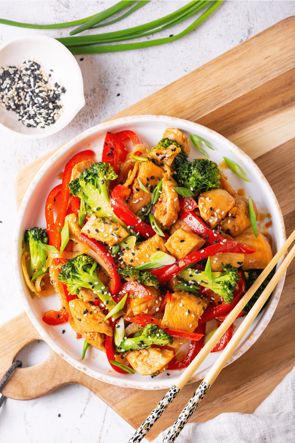 An overhead view of a white bowl filled with chicken stir fry. The bowl is on a wooden cutting board on a white table.