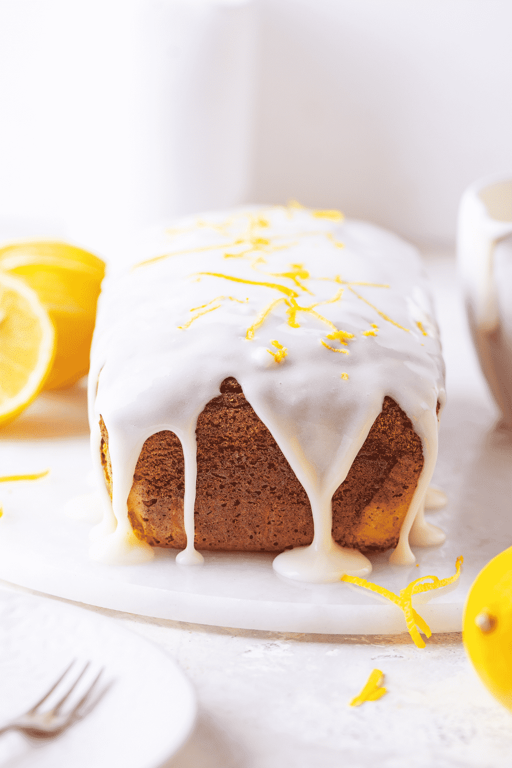 A loaf of keto lemon pound cake with keto glaze dripping down the side front of the cake. There is a lemon zest on top of the white glaze. The low carb lemon pound cake is on a white saucer on a white table.