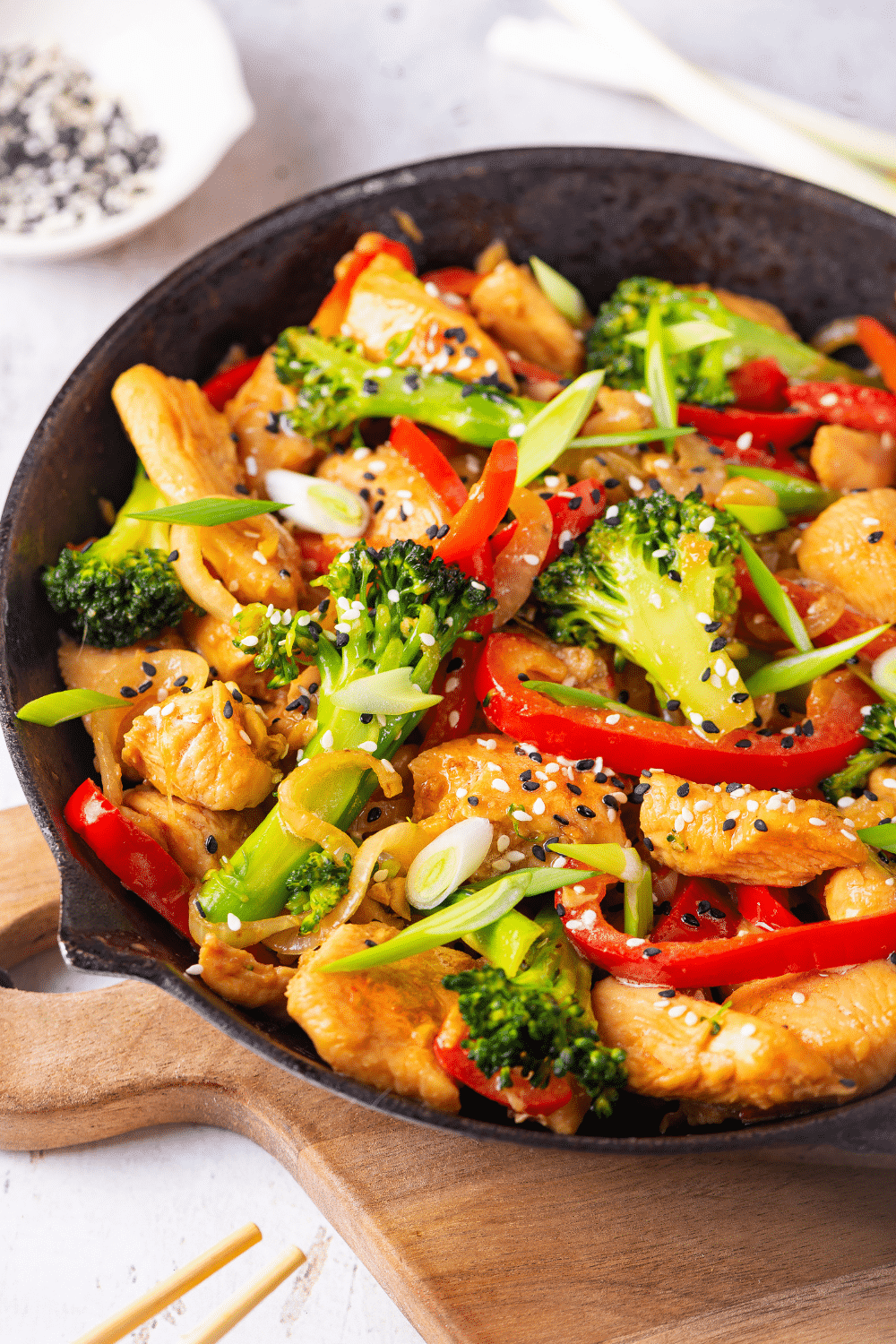 A skillet filled with keto stir fry. The skillet has chicken, broccoli, red peppers, and onions in it. Everything is covered in a keto stir fry sauce. The skillet is on a wooden cutting board on a white counter.