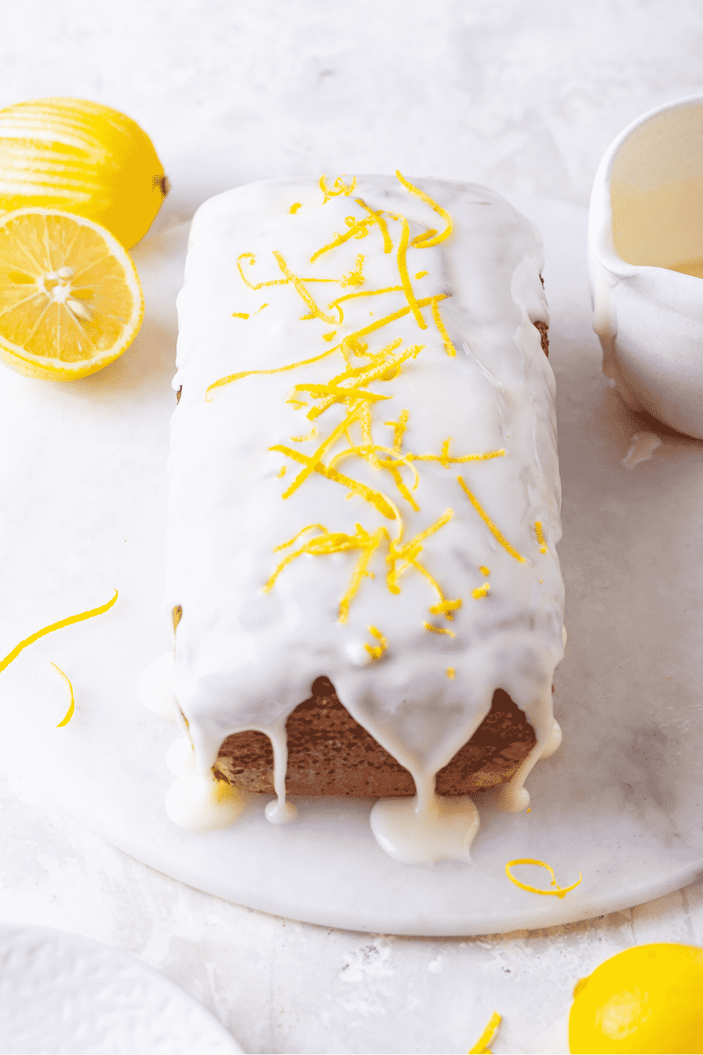 A loaf of keto lemon pound cake on a white saucer. Keto glaze is dripping down the front and side of the cake onto the saucer. A lemon zest is on top of the cake on the keto glaze. There are two lemons to the back left of the cake and a bowl of keto glaze to the right.