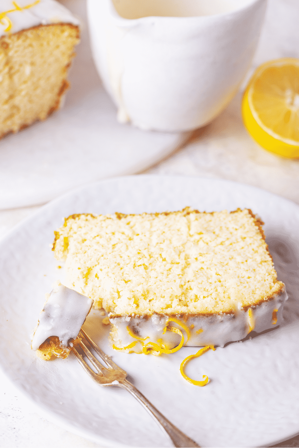 A slice of keto lemon pound cake on a white plate with top of the cake at the front of the plate. A fork has a piece of the keto lemon pound cake on it with the keto glaze facing up.