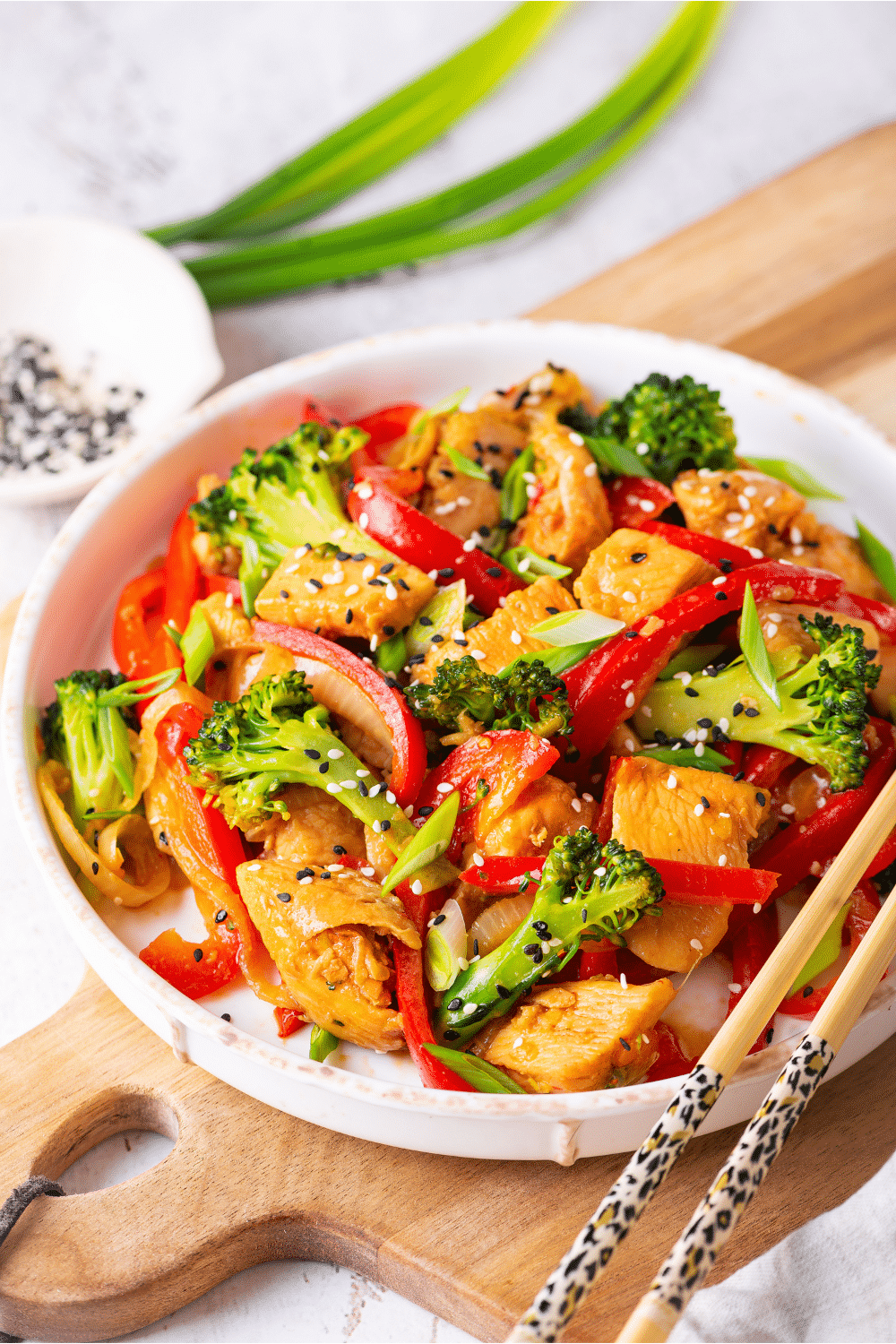 A white bowl filled with keto chicken stir fry. The bowl is on a wooden cutting board on a white table. Chopsticks are at the edge of the bowl.