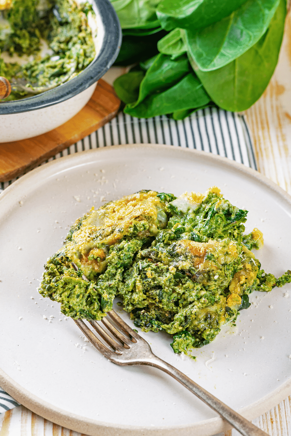 A white plate with a scoop of spinach casserole on it. A fork is rusting on the plate with some spinach casserole in the prongs.