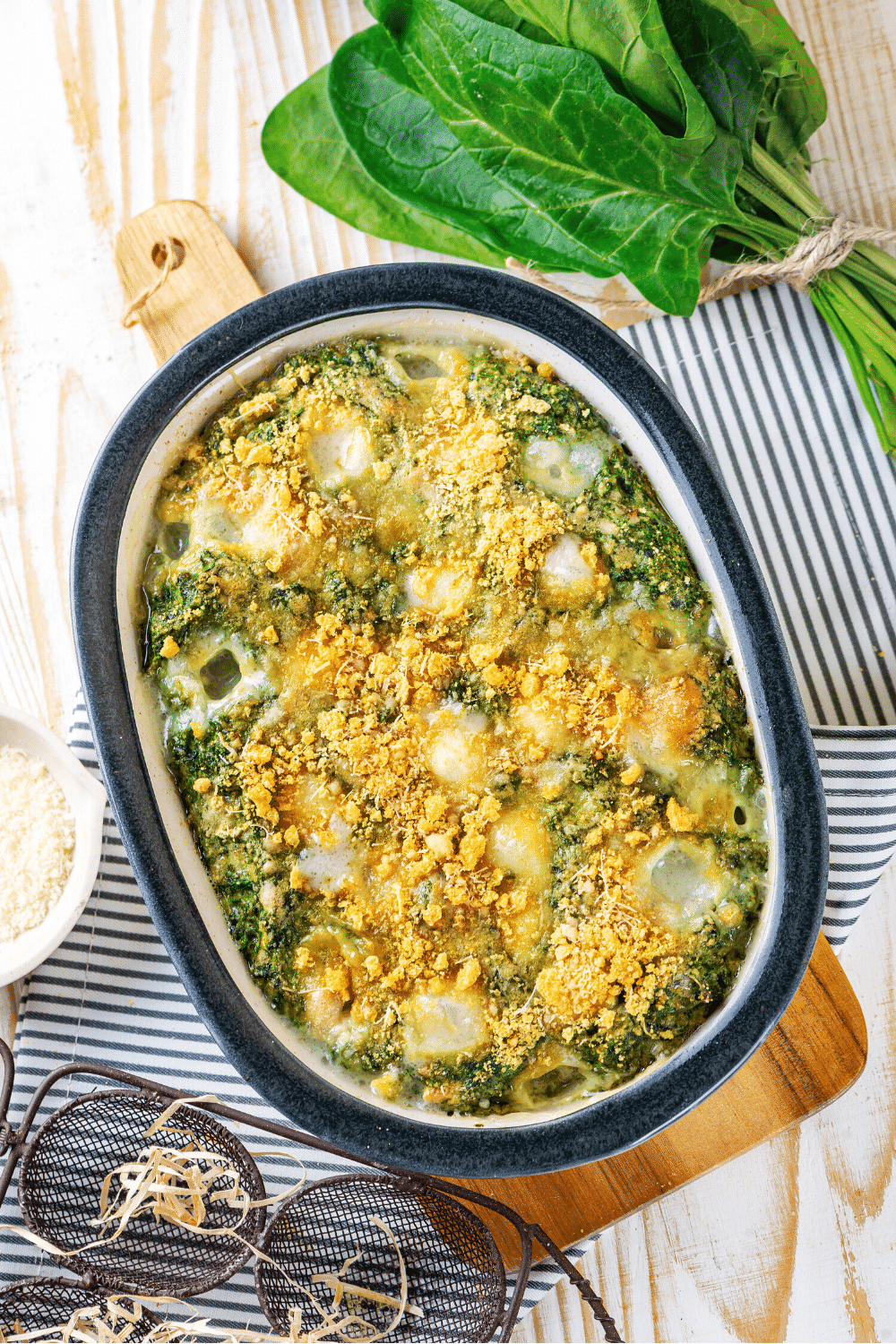A baking dish filled with cheesy spinach casserole. The baking dish is on a wooden cutting board on a white and blue stripped tablecloth. A bundle of spinach is to the back right of the spinach casserole.