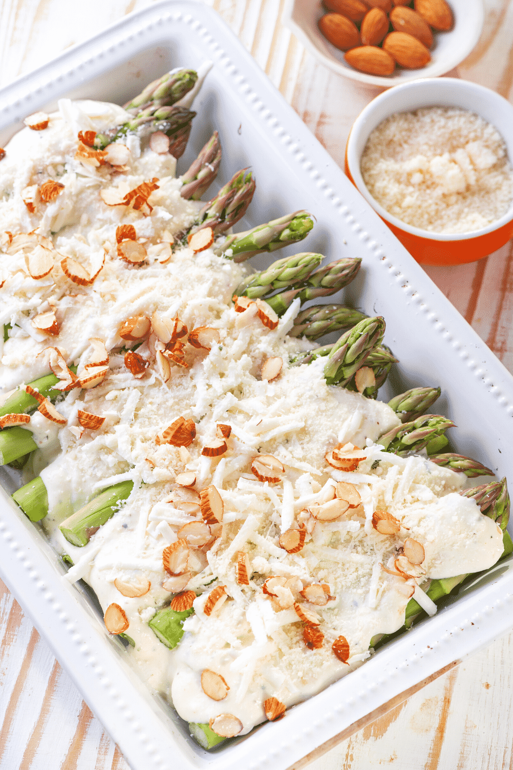 A white casserole dish filled with asparagus topped with a cream sauce, cheese, and sliced almonds.