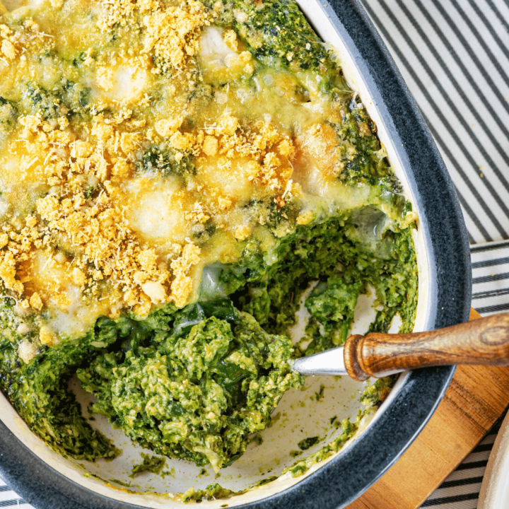 A baking dish filled with cheesy spinach casserole. A spoon is scooping out some spinach casserole from the front of the baking dish