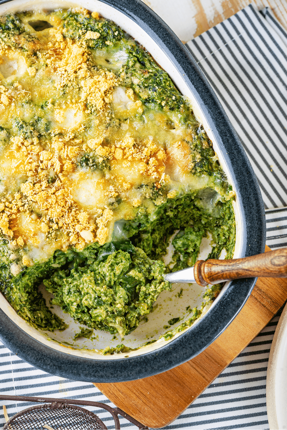 A baking dish filled with cheesy spinach casserole. A spoon is scooping out some spinach casserole from the front of the baking dish