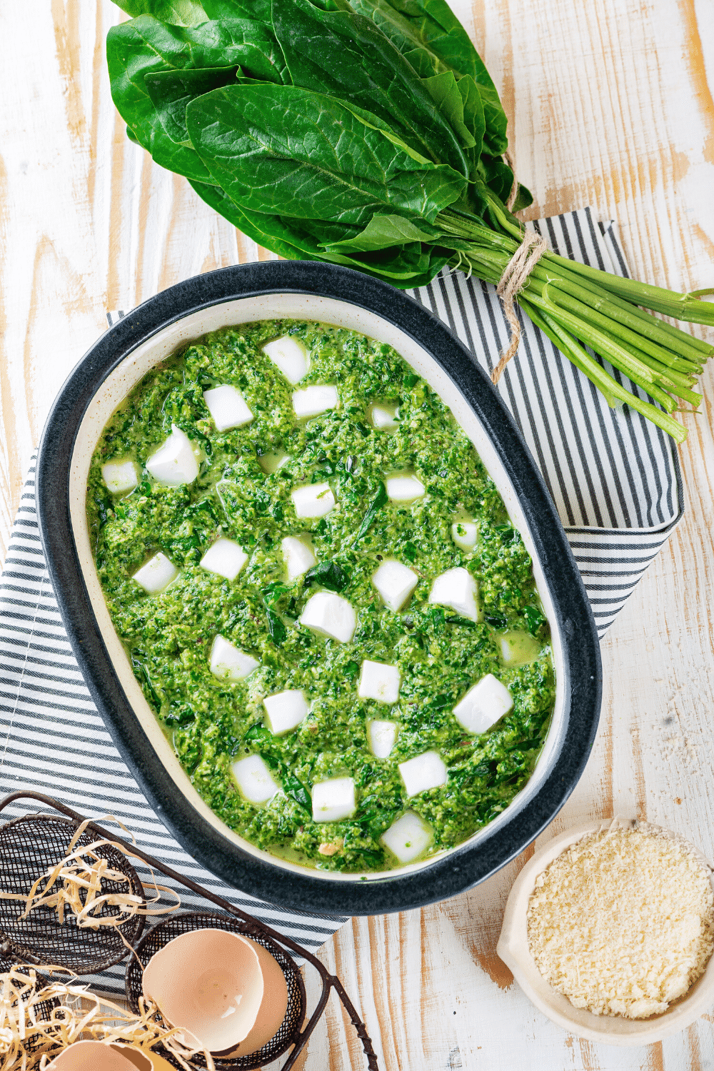 A baking dish with spinach casserole and feta cheese cubes on top. There is a bundle of spinach behind the baking dish and they are on a white and blue stripped tablecloth. A small bowl of breadcrumbs is on the wooden table in front the spinach casserole.