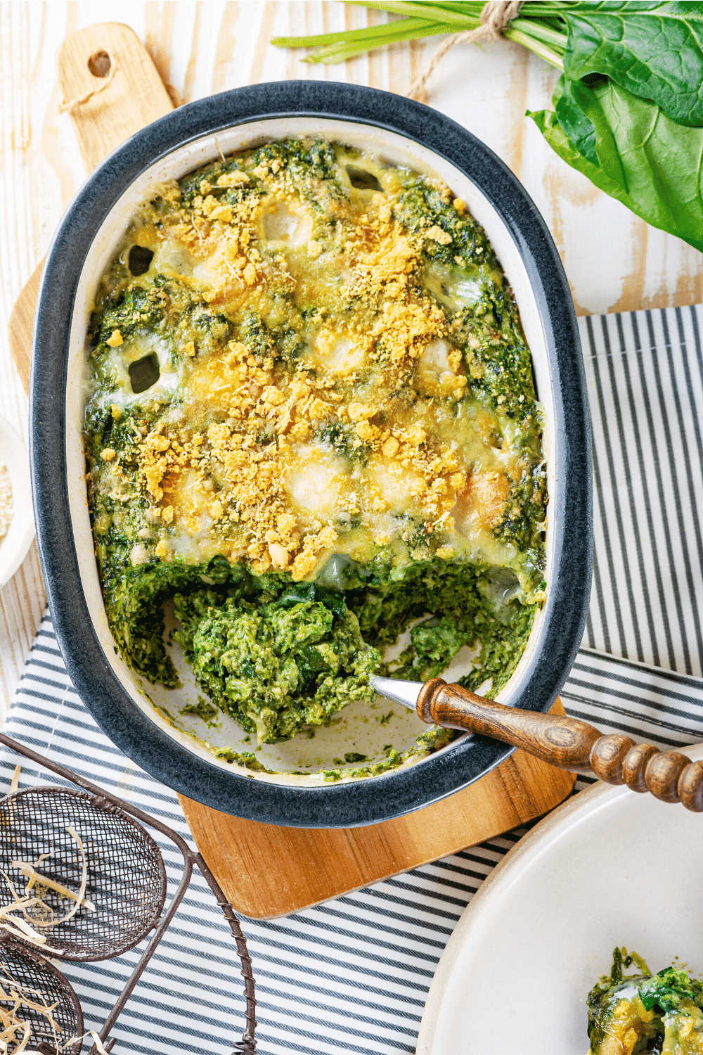 A baking dish filled with spinach casserole. A spoon is scooping up some of the spinach casserole. The baking dish of spinach casserole is on a wooden cutting board on top of a white and blue tablecloth.