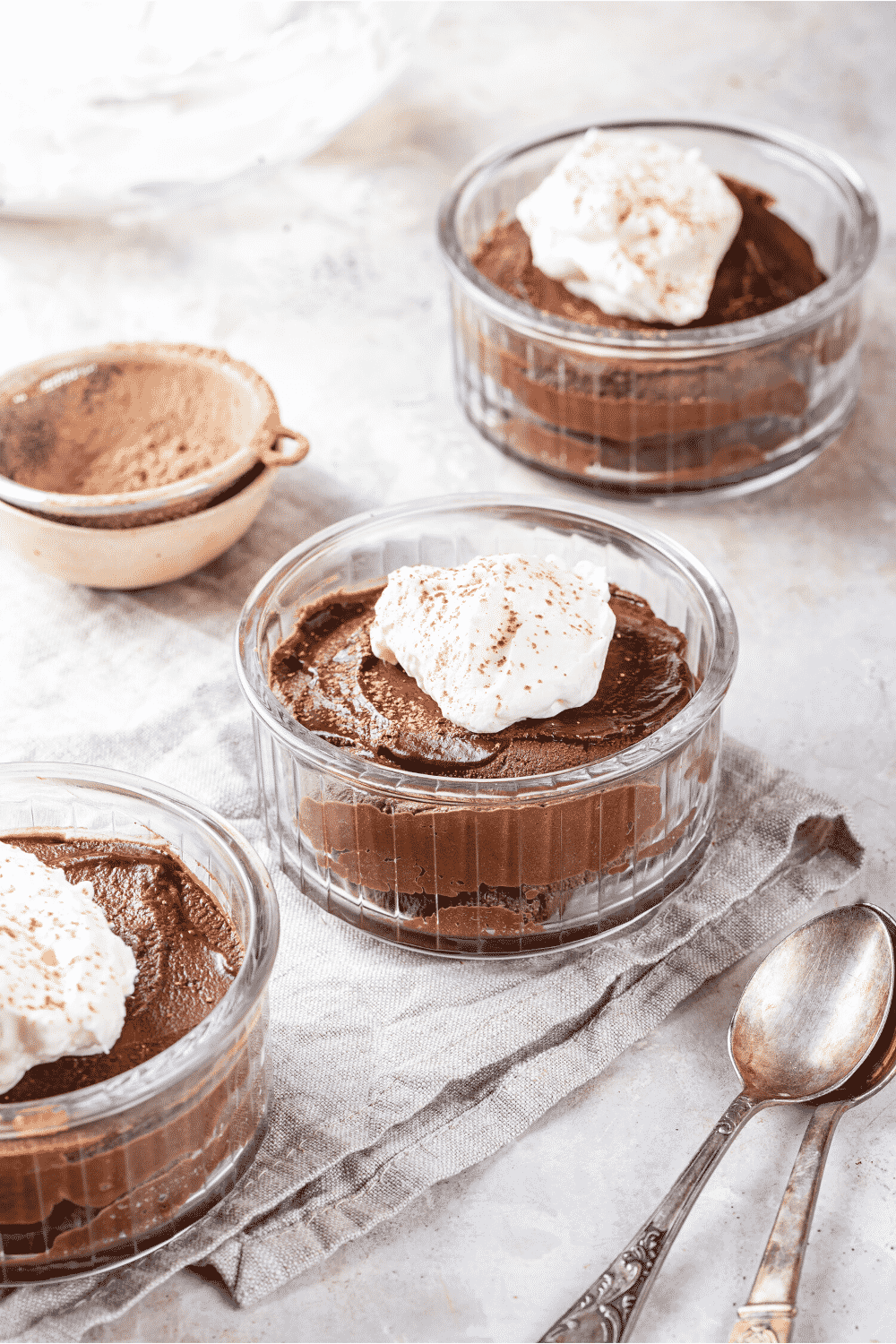 Three glass bowls of keto chocolate mousse lined up unevenly in a row. There is whipped cream on top of the keto mousse and the bowls are on a grey tablecloth. To the left of the bowl in the middle is a small bowl of cocoa powder.