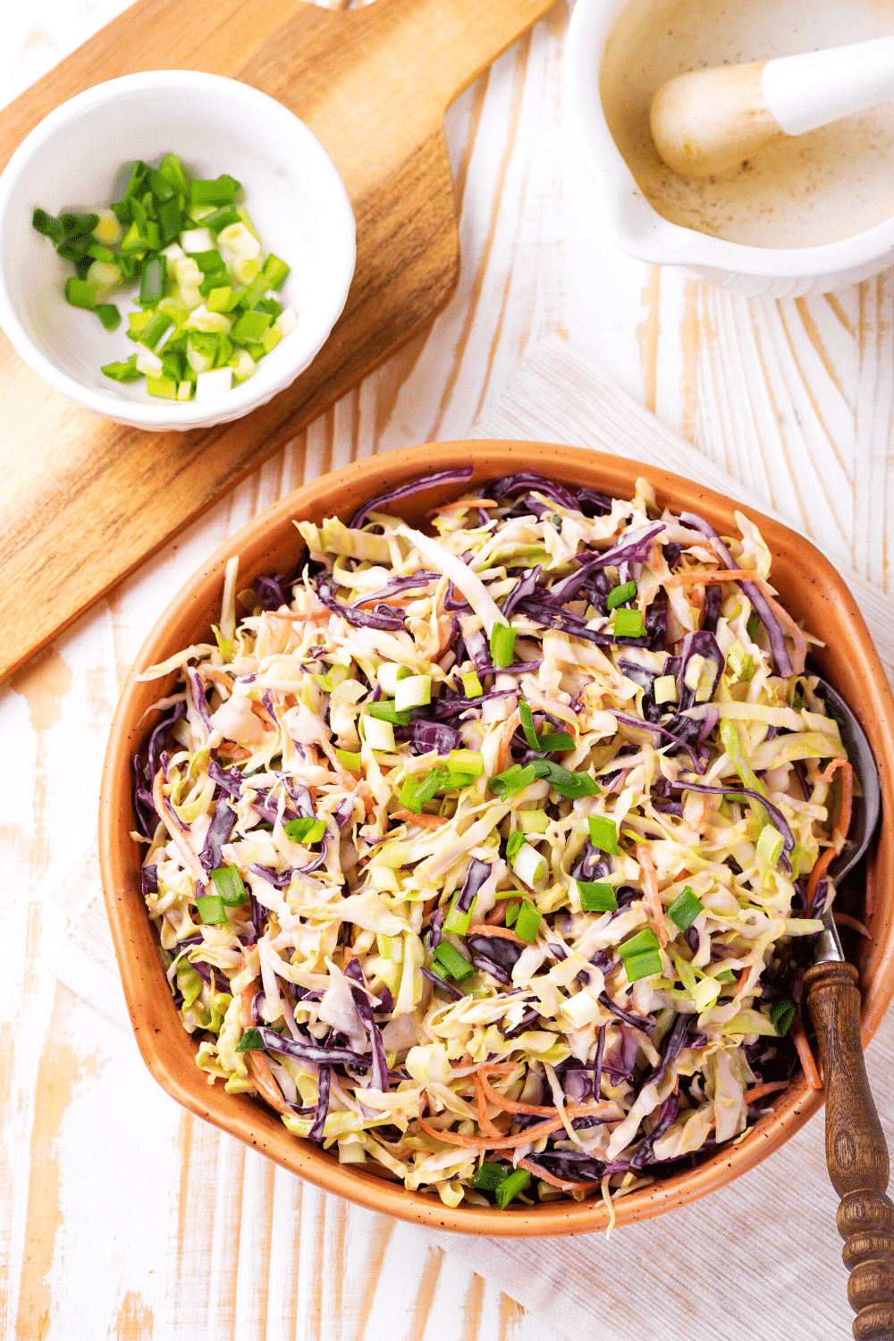 A wooden bow filled with keto coleslaw. The bowl is on a white tablecloth on top of a wooden table. Behind the bowl is a wooden cutting board with a white bowl filled with chives.