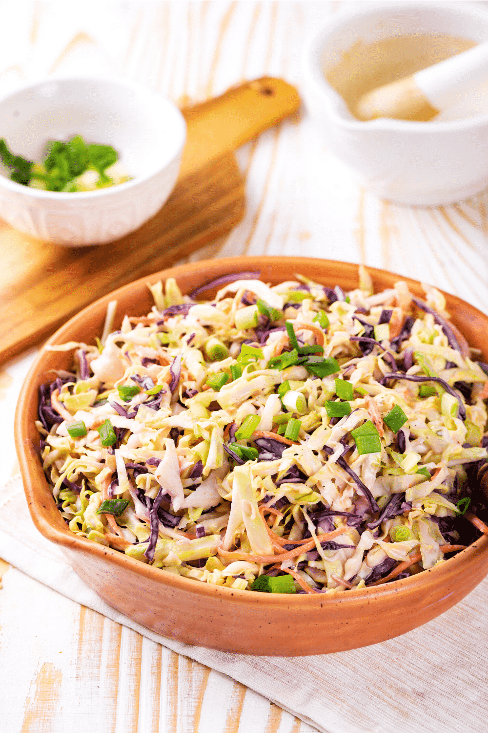 A wooden bowl filled with keto coleslaw. The bowl is on white tablecloth on top of a wooden table. A wooden cutting board with a white bowl of chives is behind the bowl of low carb coleslaw.