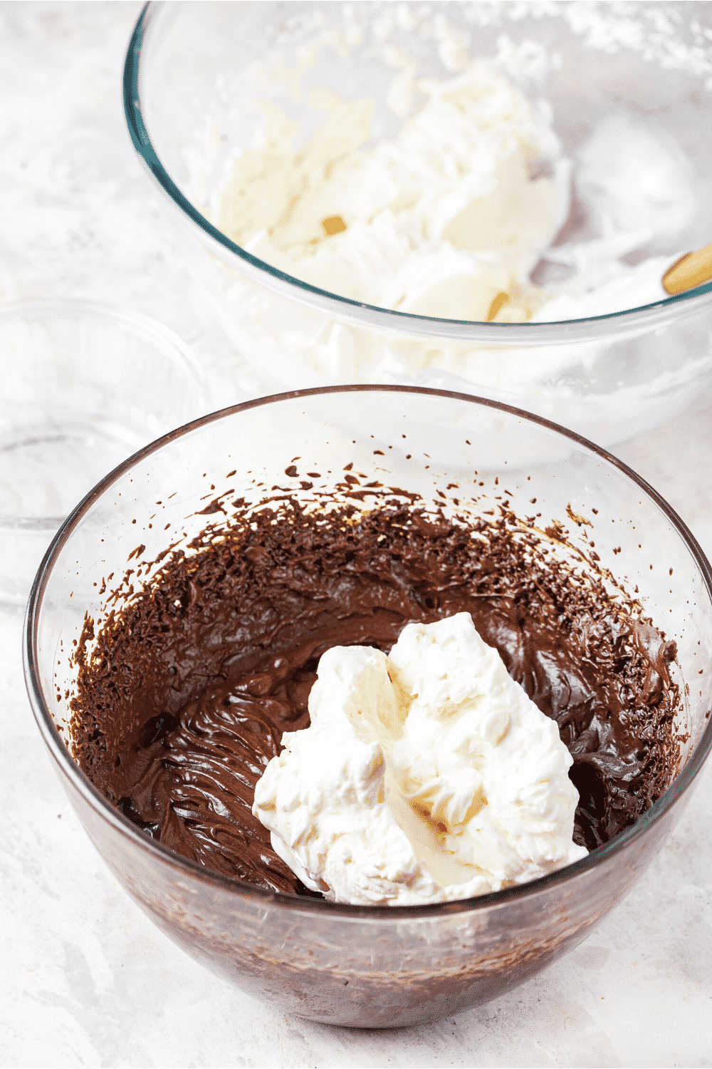 A glass bowl of whipped keto chocolate with some whipped cream on top. There is a glass bowl of whipped cream behind the keto mousse with a rubber spatula in the bowl.