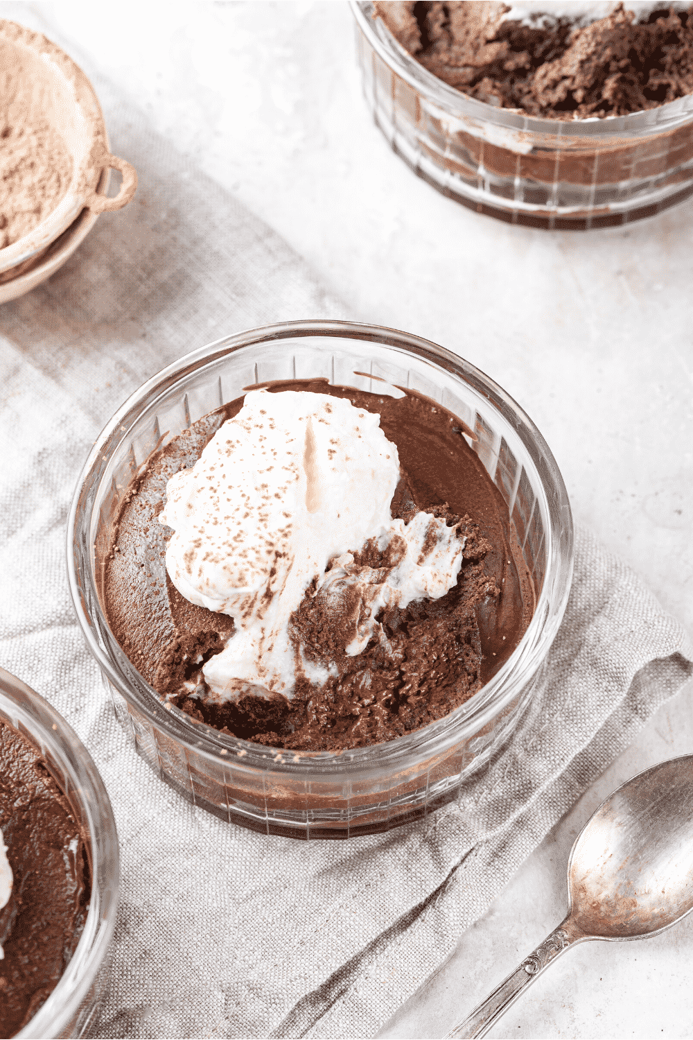 A glass bowl filled with keto chocolate mousse. Keto whipped cream is on top of the mousse and the glass bowl is on a grey tablecloth.