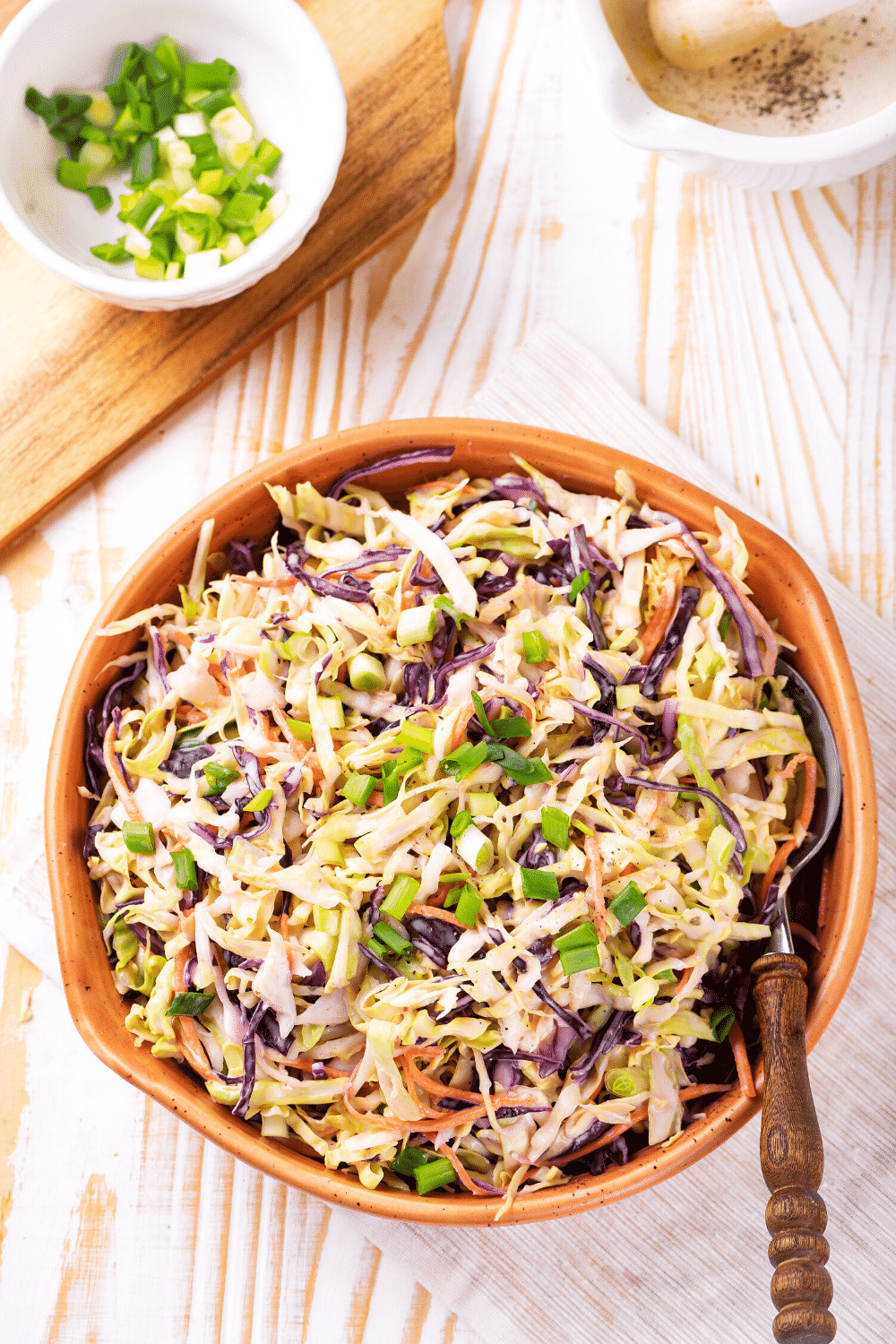 An overhead view of keto coleslaw in a wooden bowl. A spoon is emerged in the keto coleslaw and the bowl is on a white tablecloth on a wooden counter.