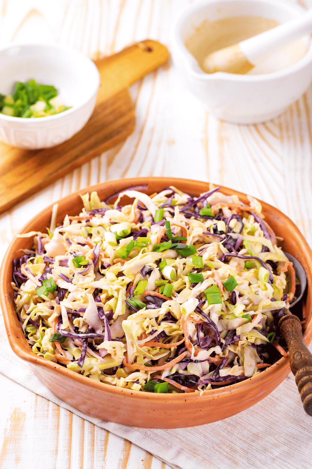 A wooden bowl filled low carb keto coleslaw. There is a spoon at the right side of the bowl in the keto coleslaw. The bowl is on a white tablecloth on a wooden counter.