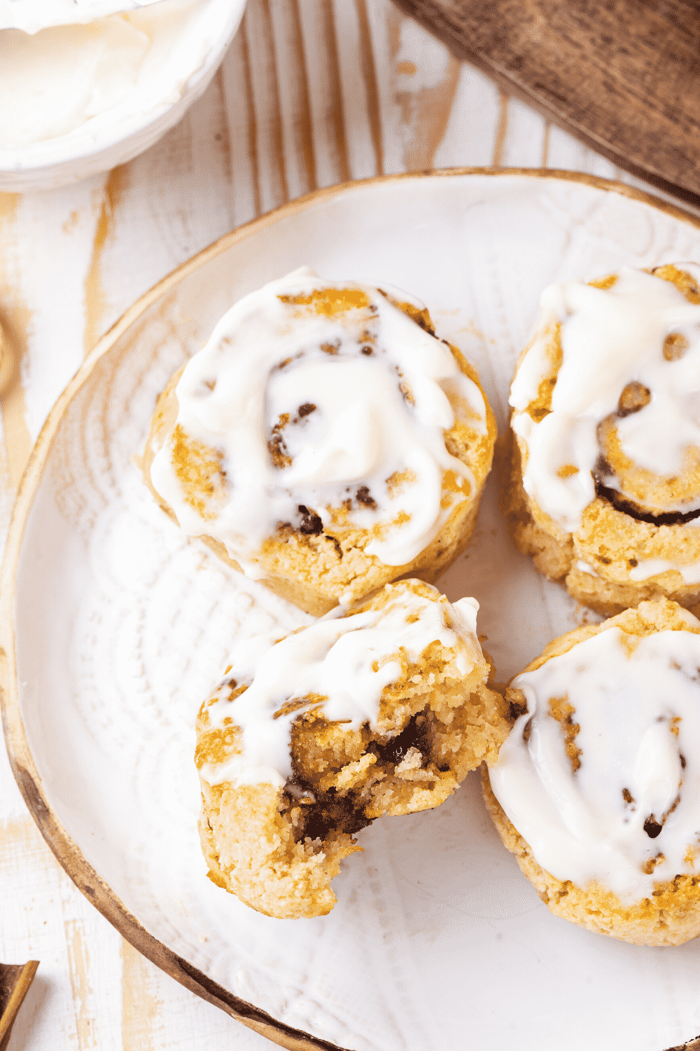 A white plate with four cinnamon rolls with icing on top. The front left cinnamon roll is on its side and has a bite out of the front.