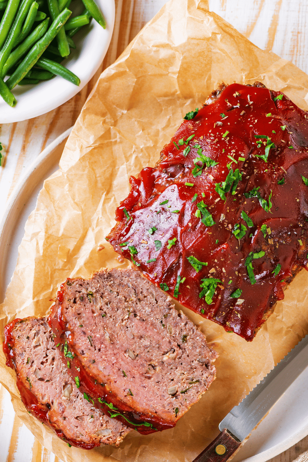 An overhead view of a whole keto meatloaf on a piece of parchment paper on a white plate. There are two slices keto meatloaf in front of the loaf.