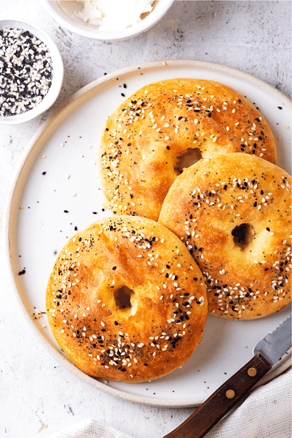 Three bagels overlapping one another on a white plate. There are small bowls of seasoning and butter behind