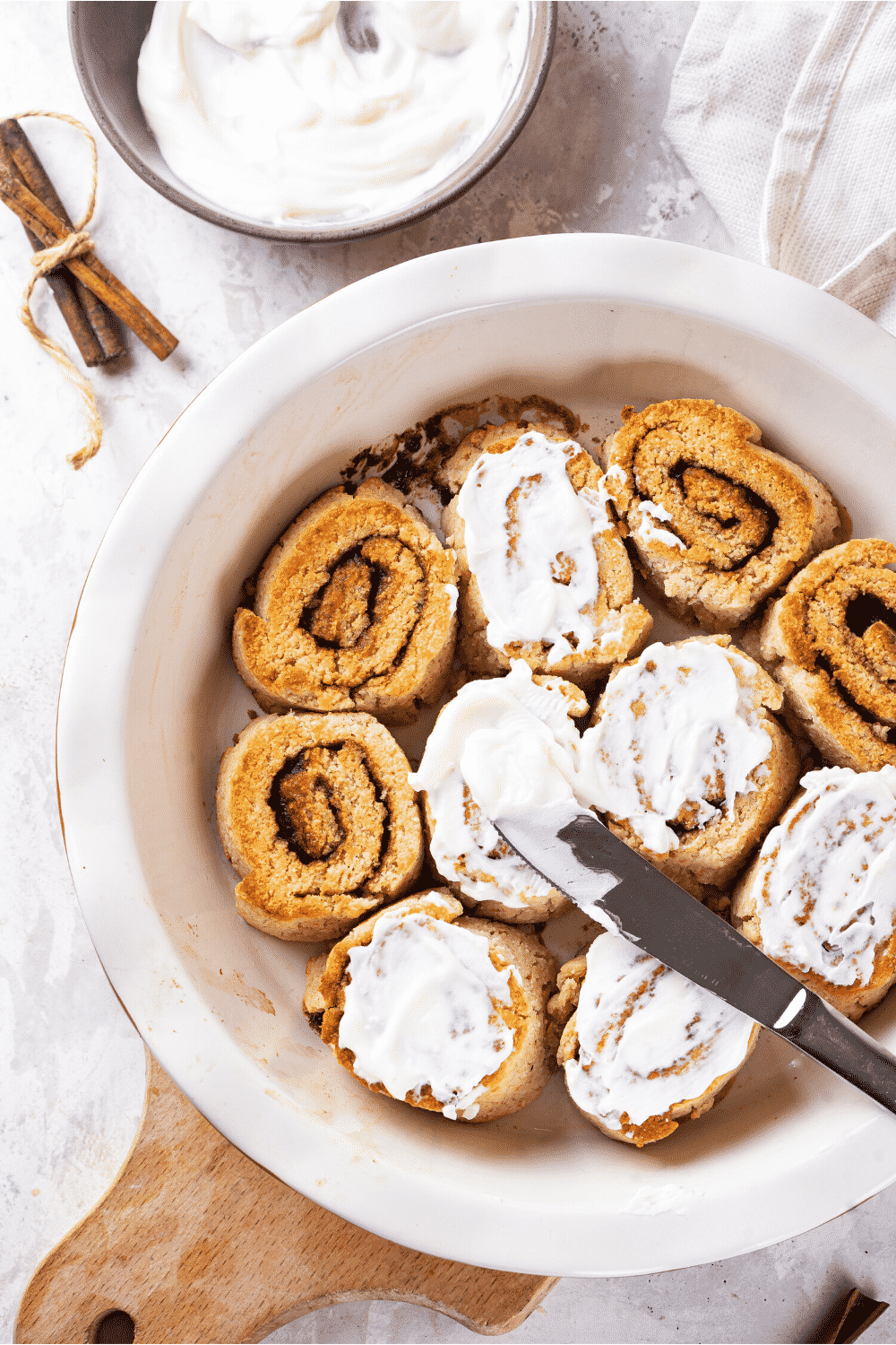 A white baking dish with 10 cinnamon rolls in it. Two cinnamon rolls at the edge of the left side and two cinnamon rolls at the edge of the right side or on frosted. All the other cinnamon rolls have icing on top and there is a bowl of icing behind the baking dish.