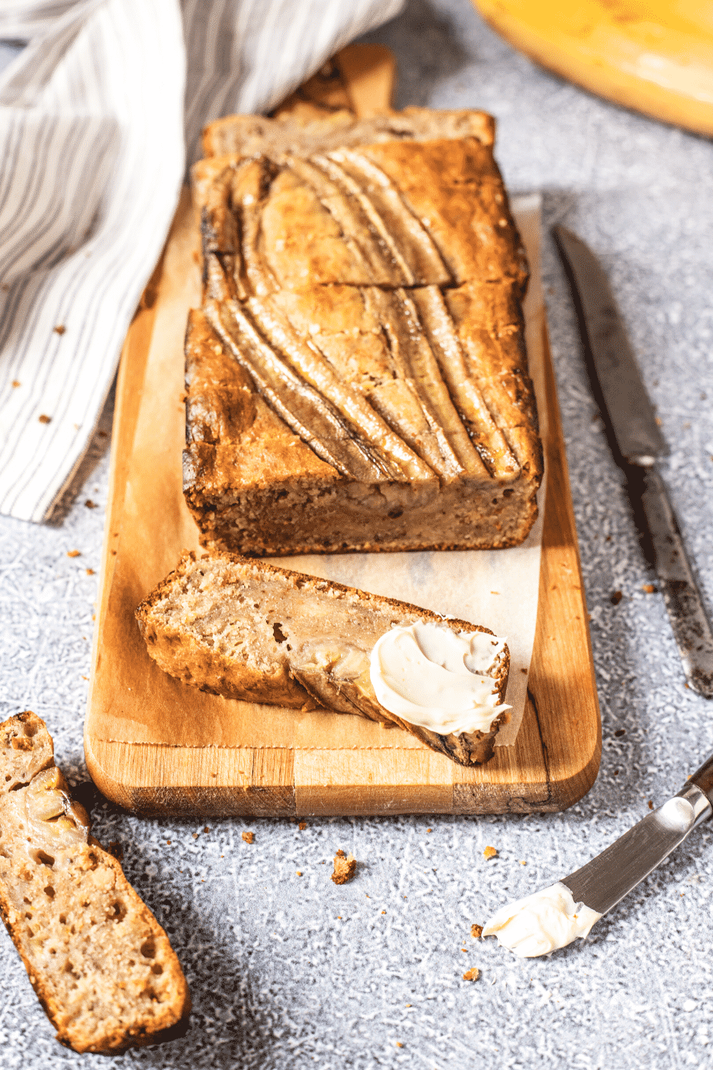 A loaf of banana bread on a piece of parchment paper on a wooden cutting board. A slice of banana bread is in front of the loaf with a slab of butter on the side. A knife is in front of the cutting board and there is butter on it.