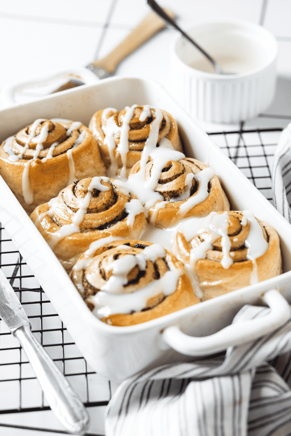 A white baking dish with six cinnamon rolls with icing on top in it. The baking dish is on a black wire rack and a cup of icing is behind the wire rack.