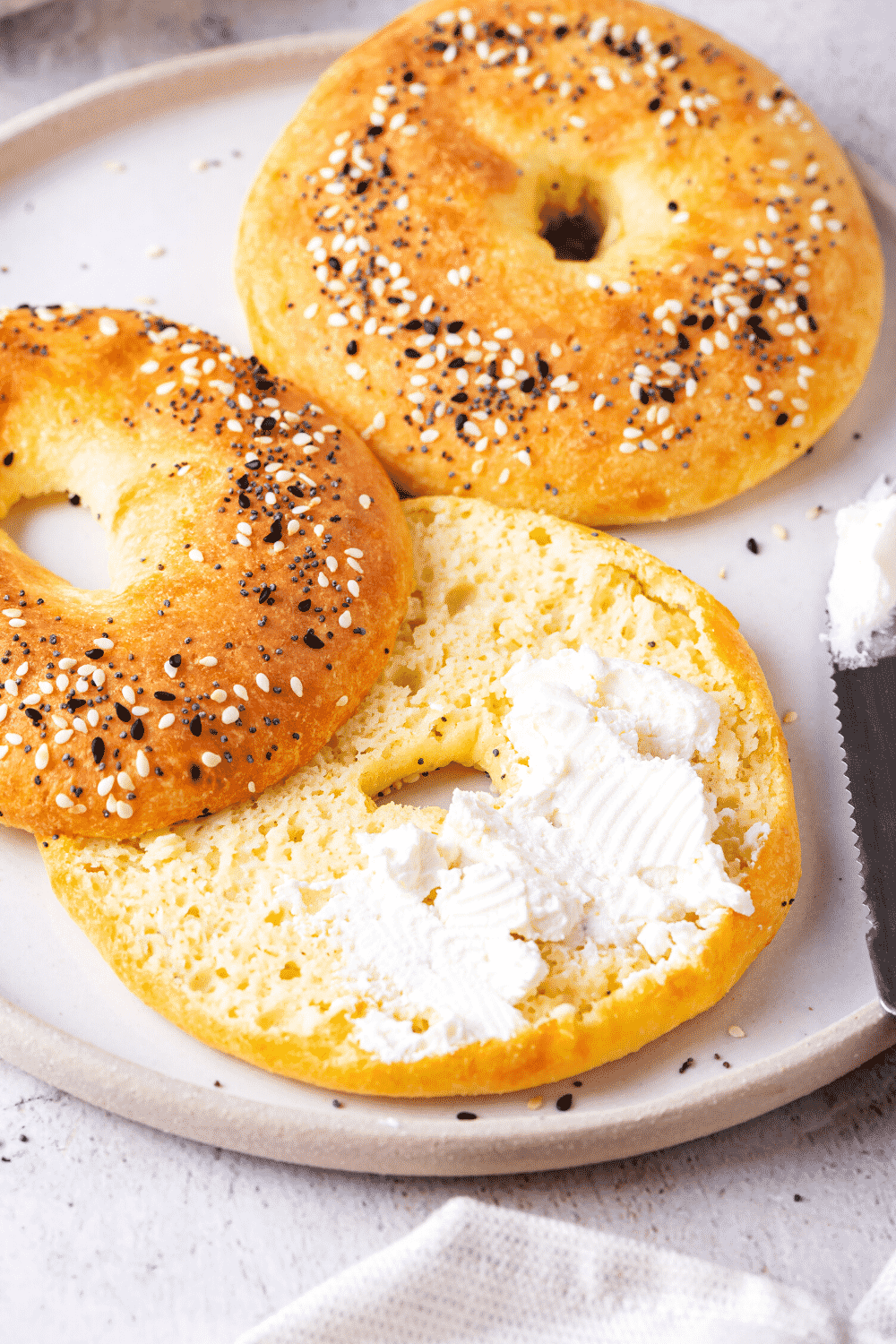 Two bagels on a white plate. A whole bagel is at the back of the plate and a sliced bagel is at the front. The sliced bagel has butter spread on the bottom half of it.