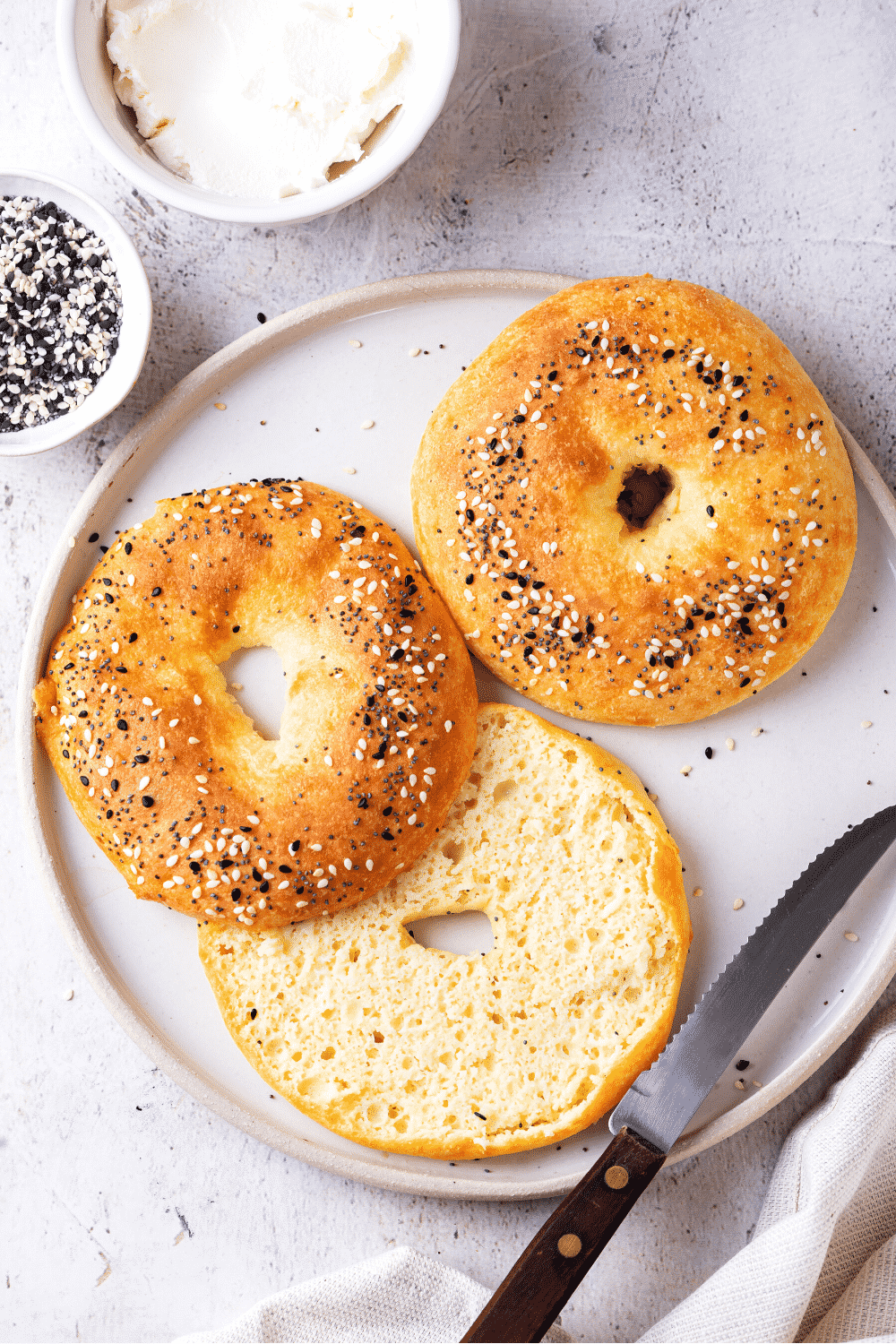 A white plate with a whole bagel and another sliced bagel next to it. A serrated knife is at the front edge of the plate.