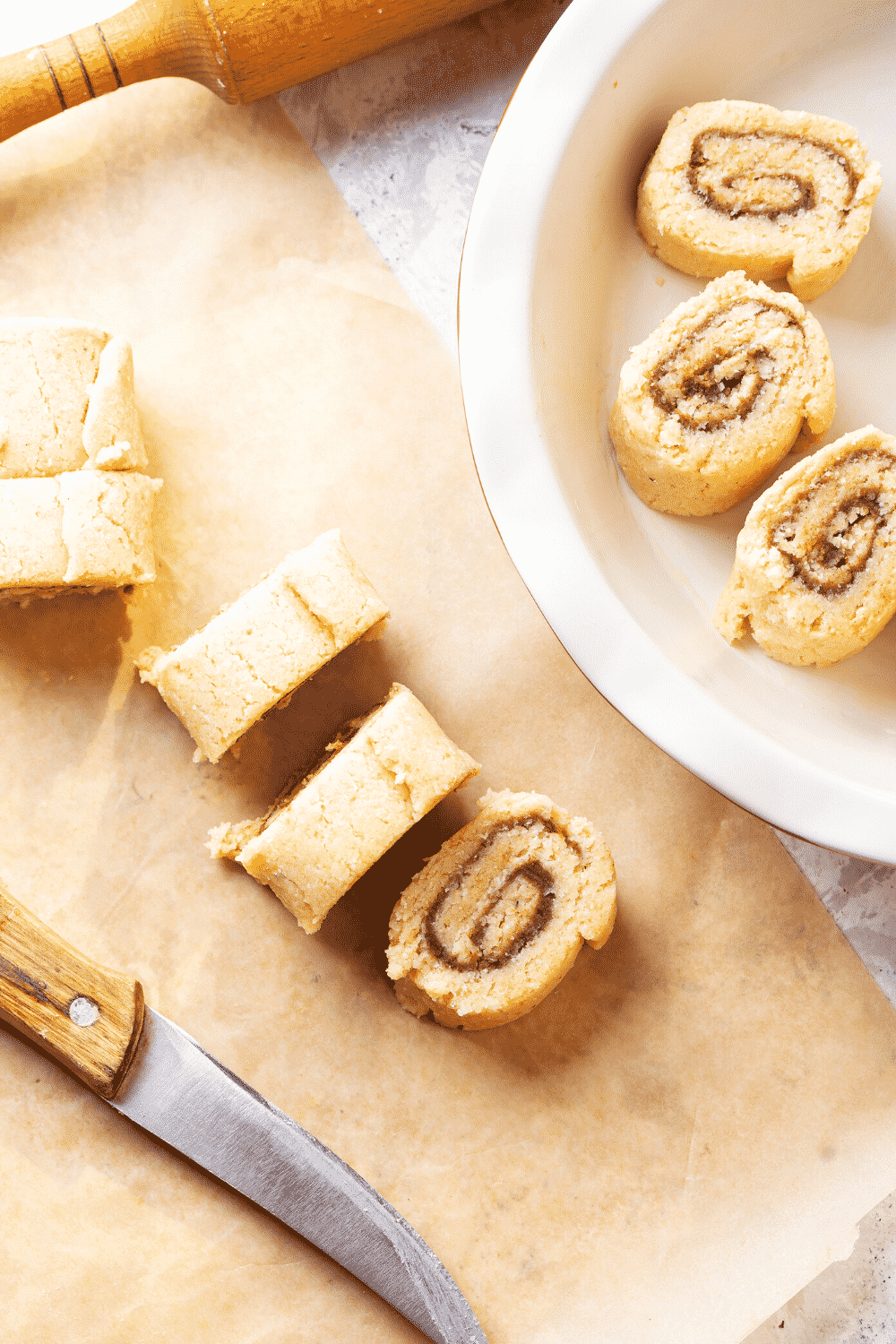 A roll of cinnamon rolls cut into five individual cinnamon rolls on a piece of parchment paper. Three other cut cinnamon rolls are in a white baking dish to the right of the piece of parchment paper.